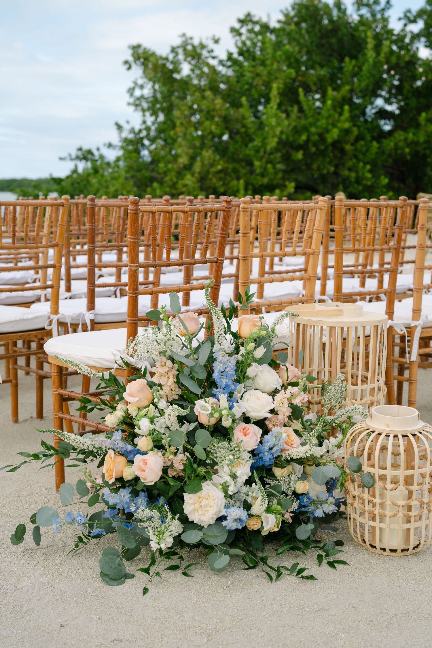Elegant floral arrangements with peach and blue flowers line wooden chairs at an outdoor wedding ceremony setup.