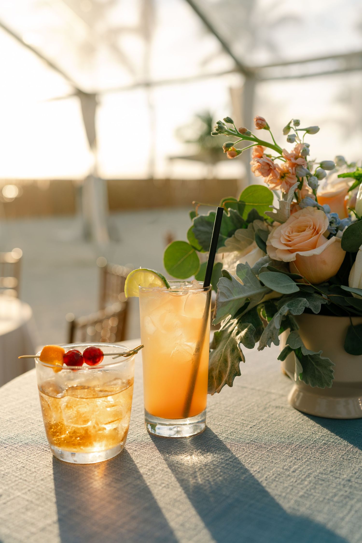 Elegant cocktails garnished with flowers and fruit sit on a sunlit table with floral arrangements.