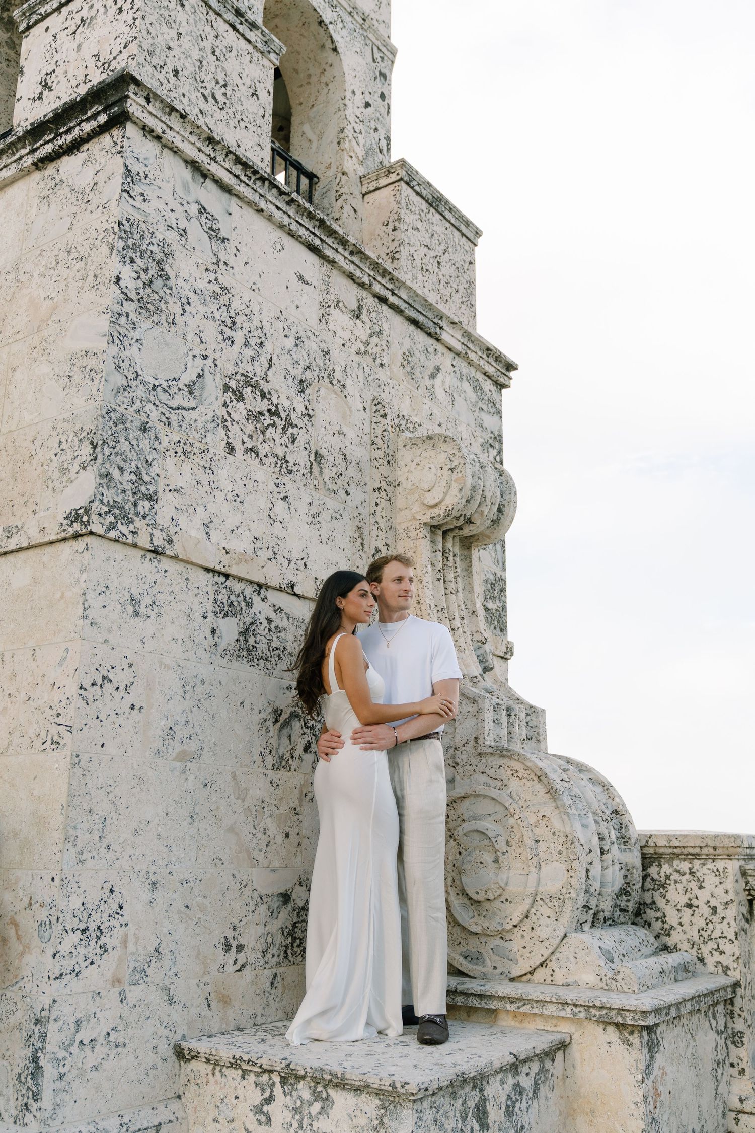 Elegant couple in white attire embracing on ornate stone steps of a historic building during a romantic photoshoot.