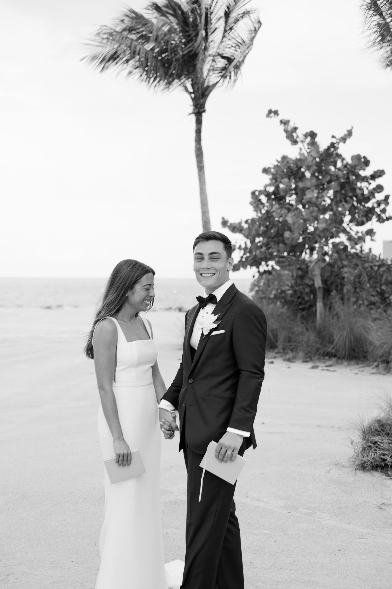 Bride and groom holding hands on a tropical beach, smiling in elegant wedding attire with palm trees behind them.