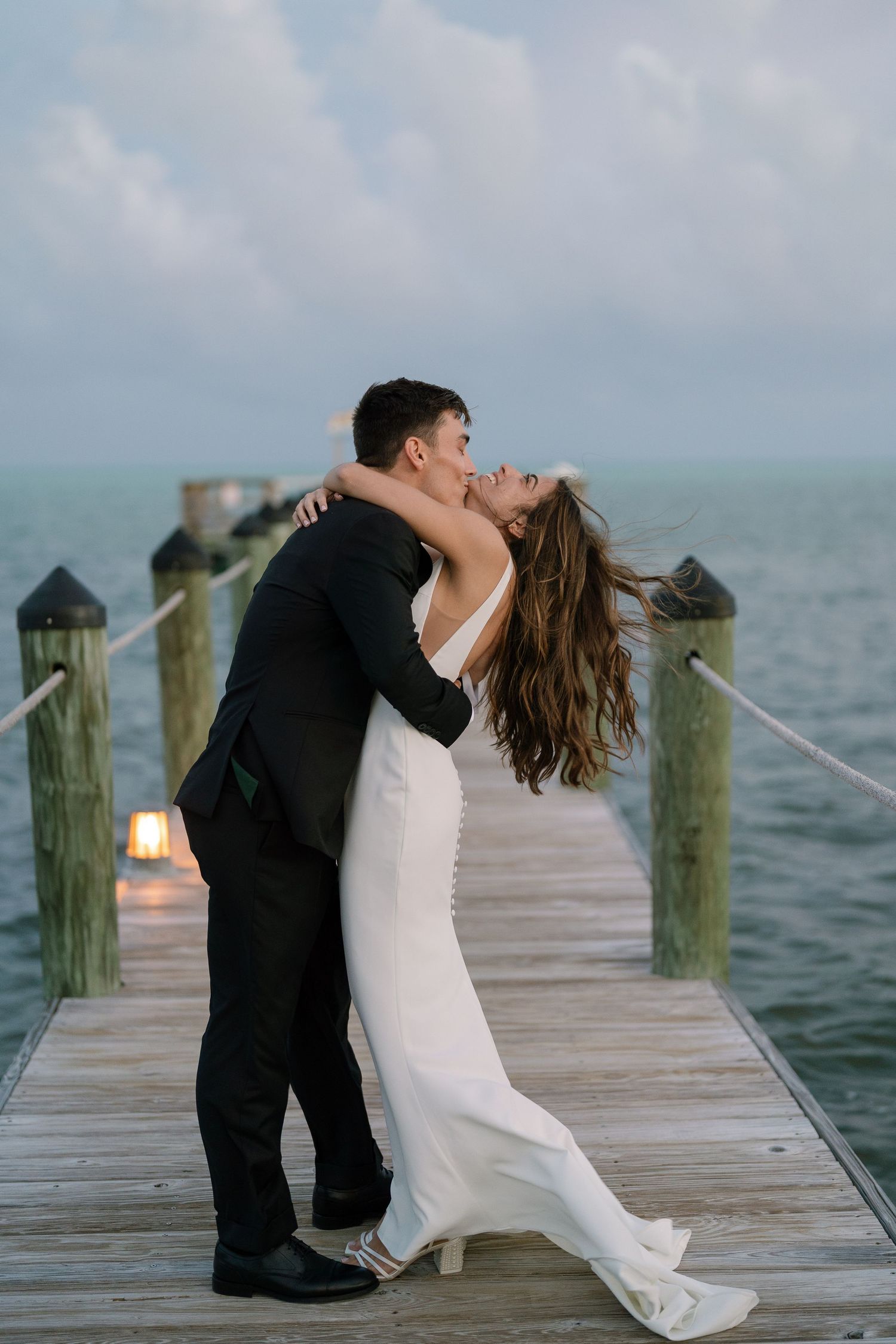 Bride and groom sharing romantic dip kiss on wooden dock over calm ocean water at dusk.
