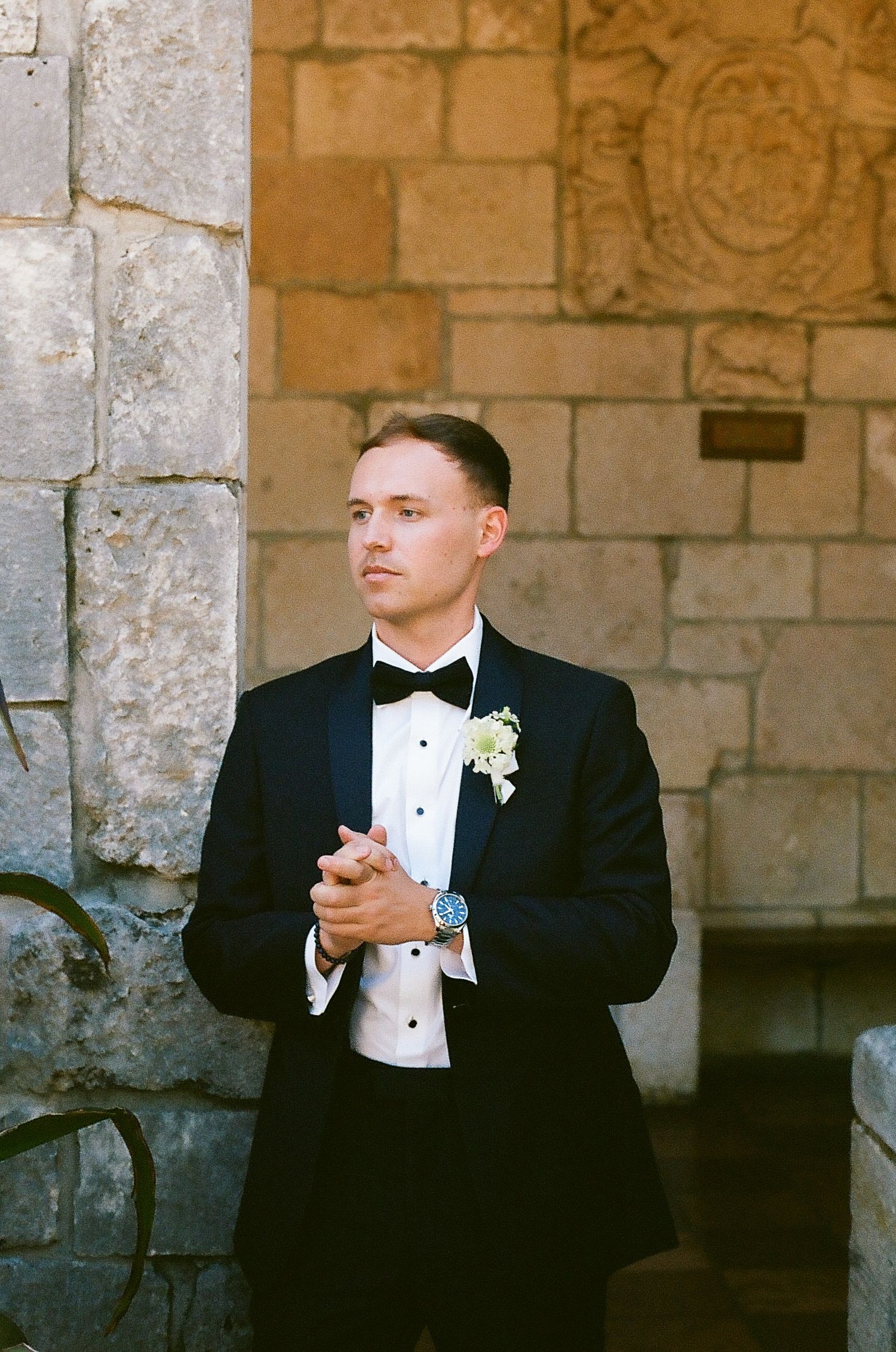 Elegant groom in black tuxedo with bow tie and boutonniere stands before stone wall, hands clasped.