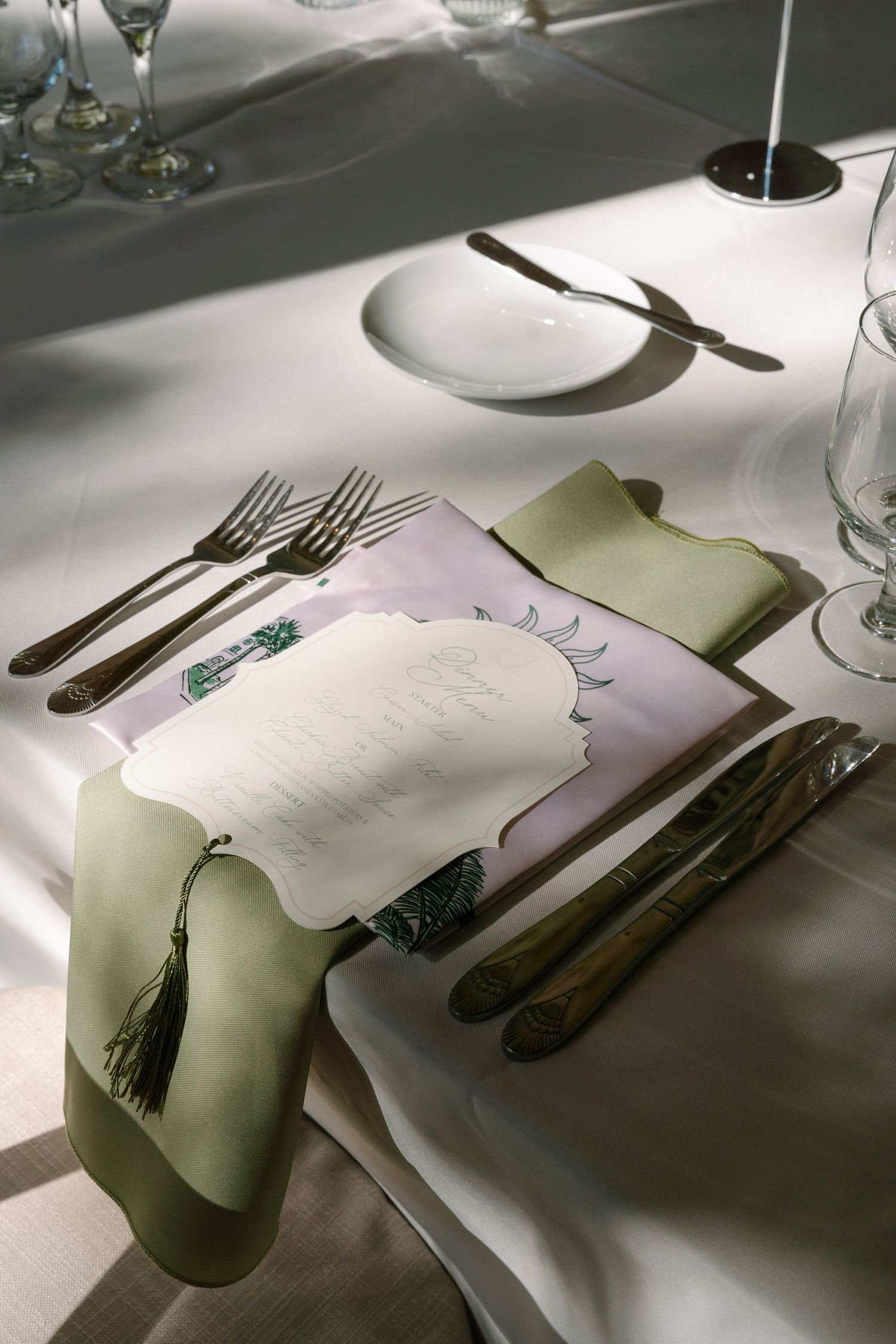 Elegant wedding place setting with green napkin, tassel, menu card, and crystal glassware on white linen.