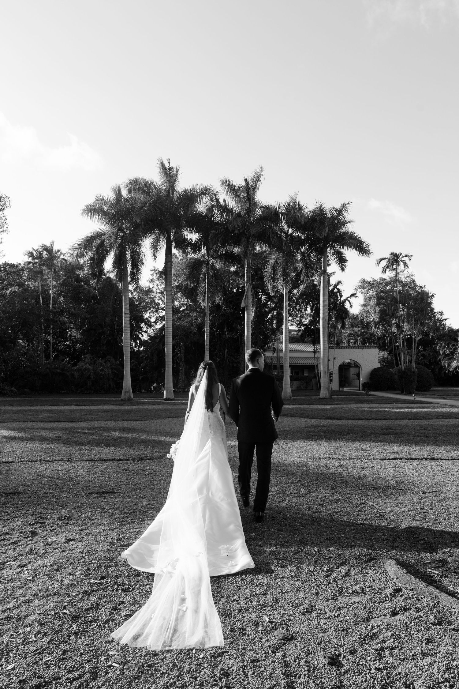 Black and white photo of bride and groom walking toward palm tree lined estate wedding venue.