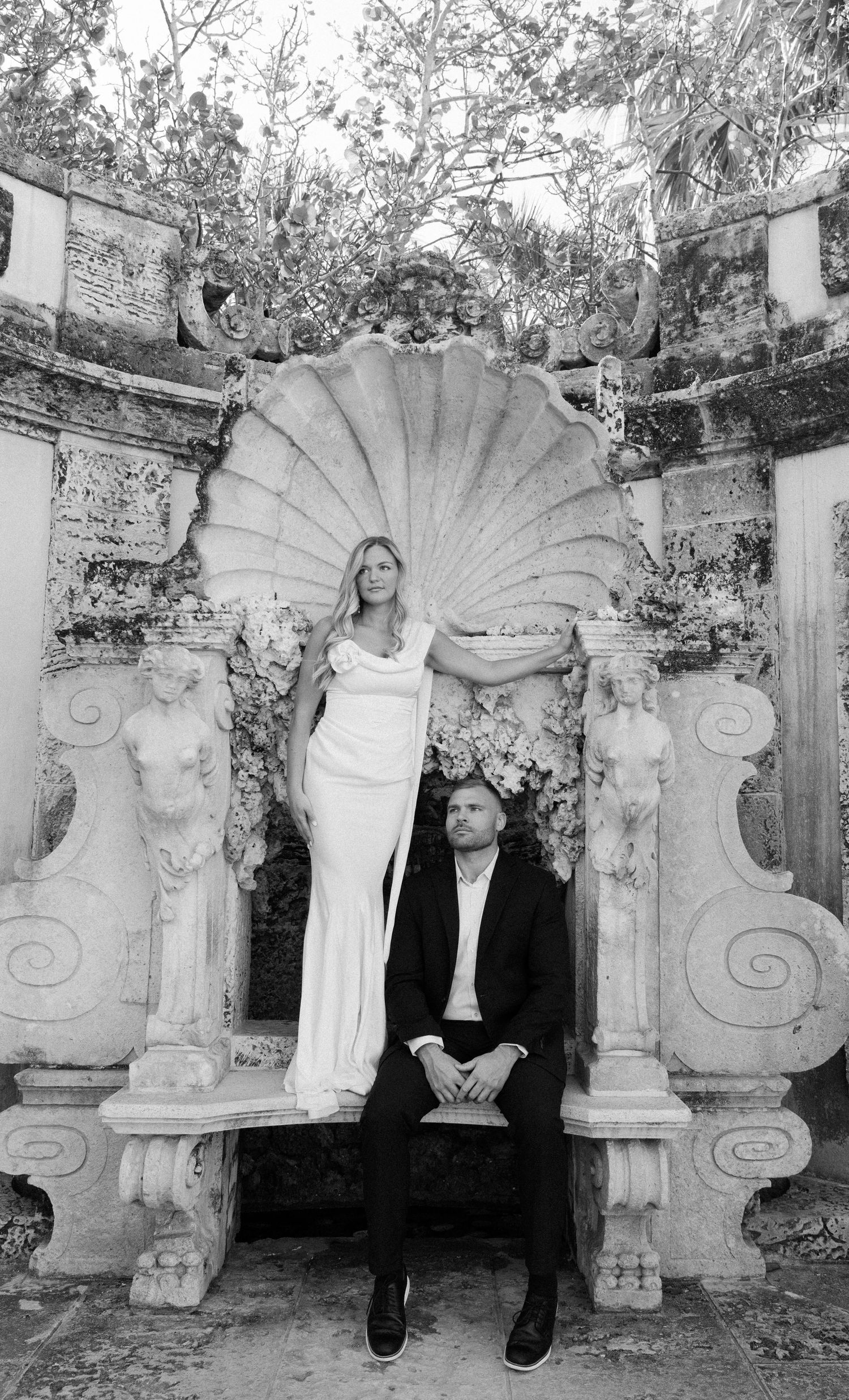 A couple poses in a classical garden setting with ornate stone architecture featuring a large shell-shaped alcove and columns.