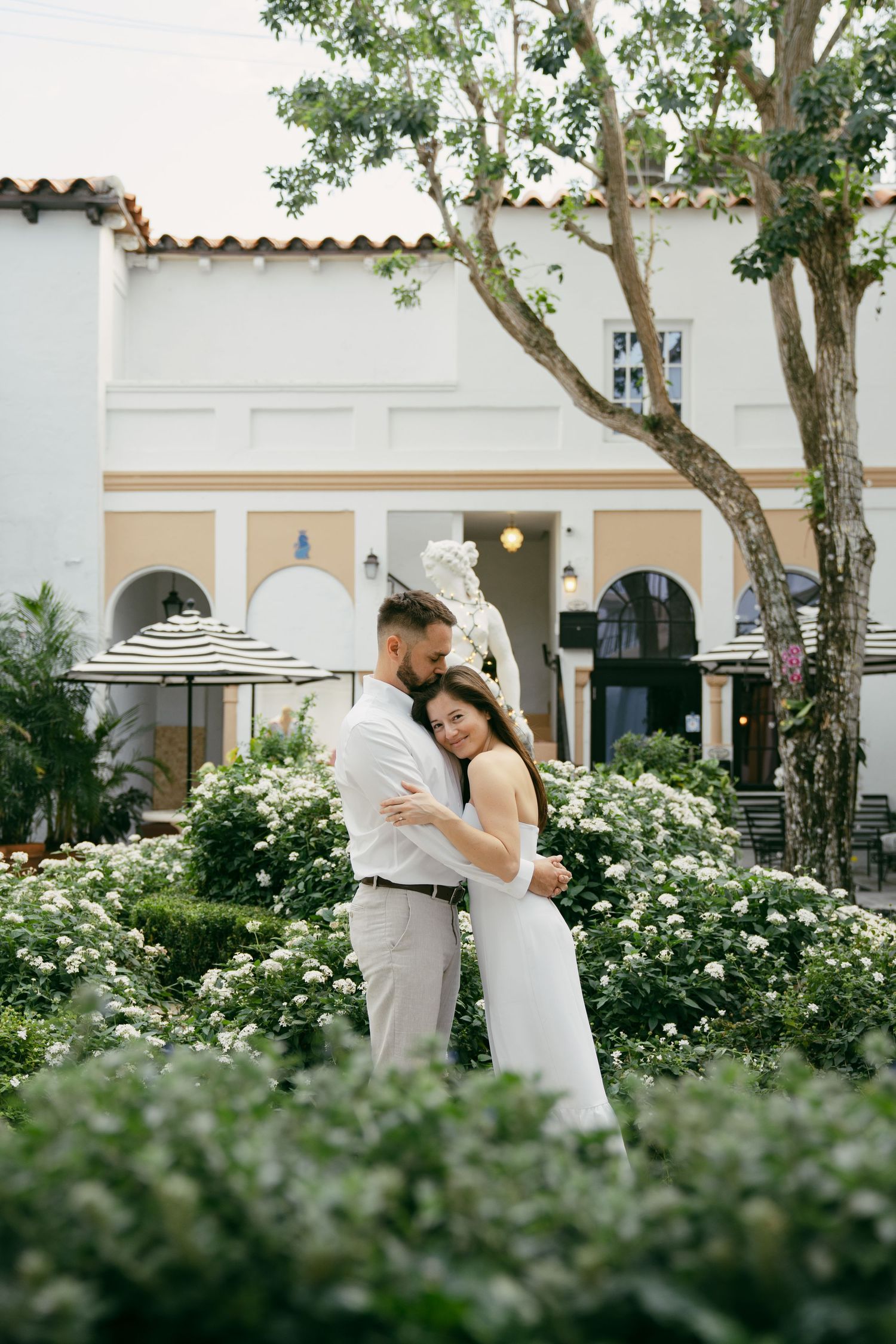 A couple embraces in a lush garden courtyard with Spanish colonial architecture and flowering bushes in the background.