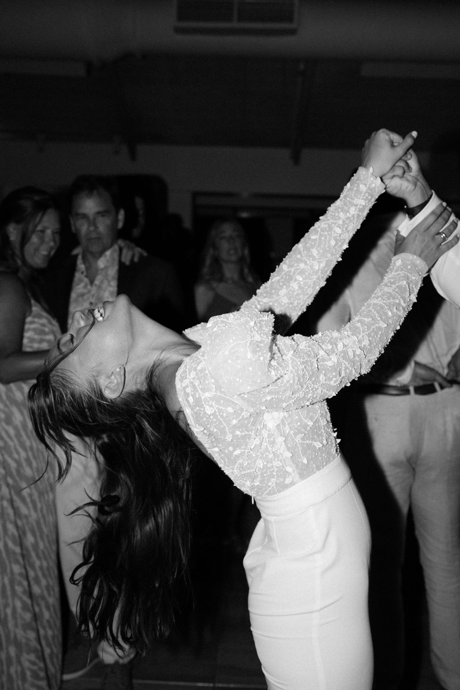 A bride in a long-sleeved white lace dress dances joyfully at her wedding reception in black and white.