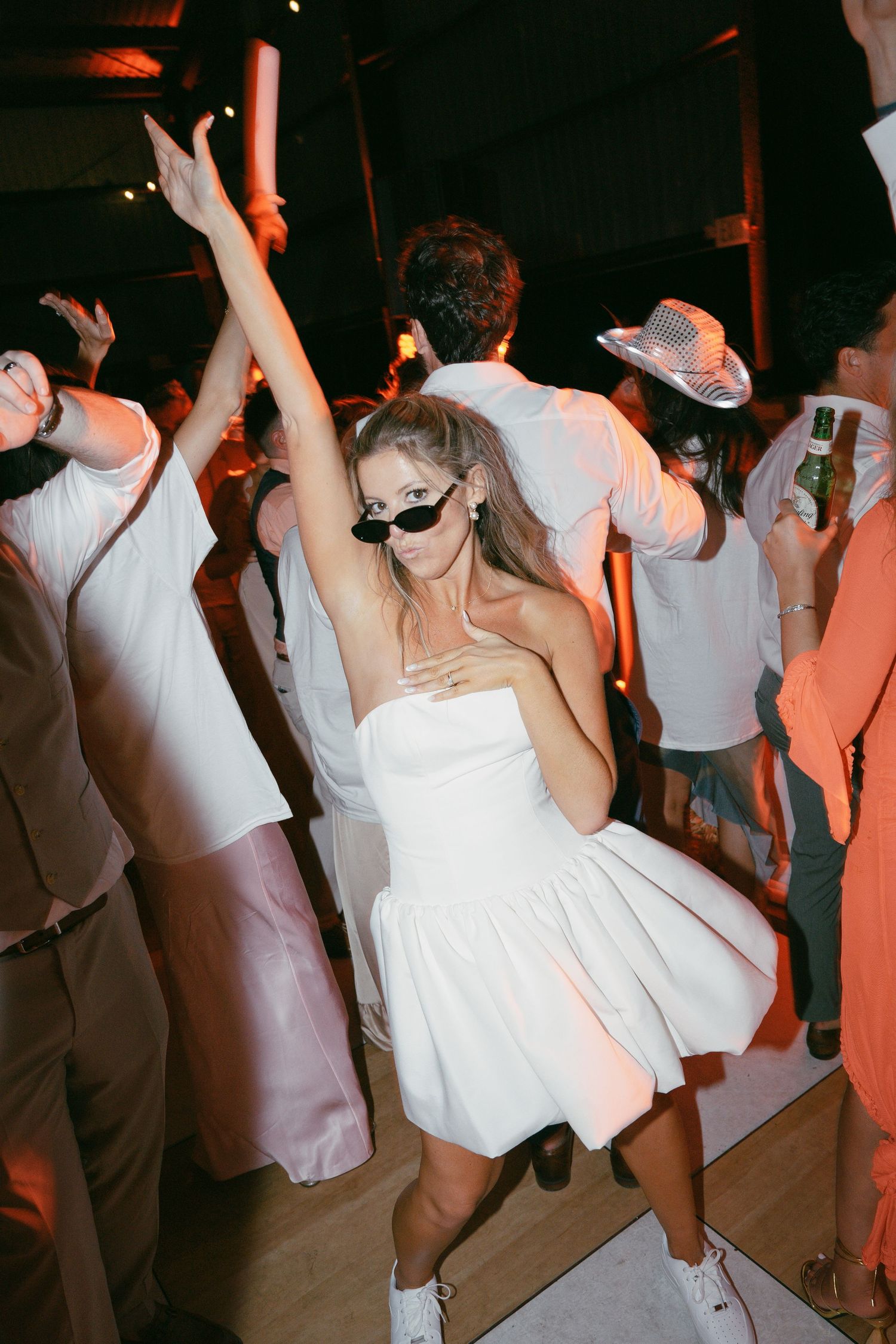 Someone in a short white dress and sunglasses dances energetically on a dark dance floor at a party.