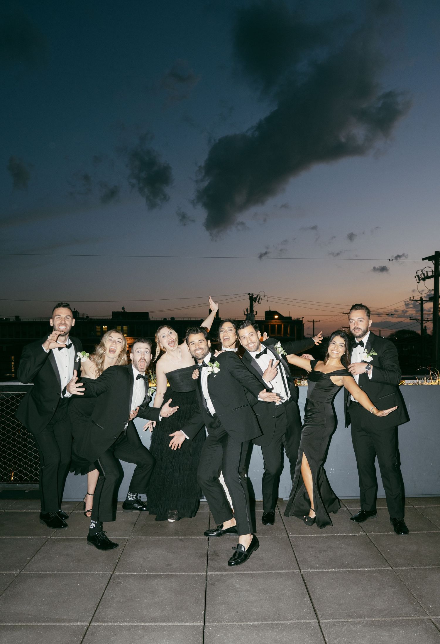 A group in formal black attire strikes playful poses on a rooftop terrace at sunset with dark clouds overhead.