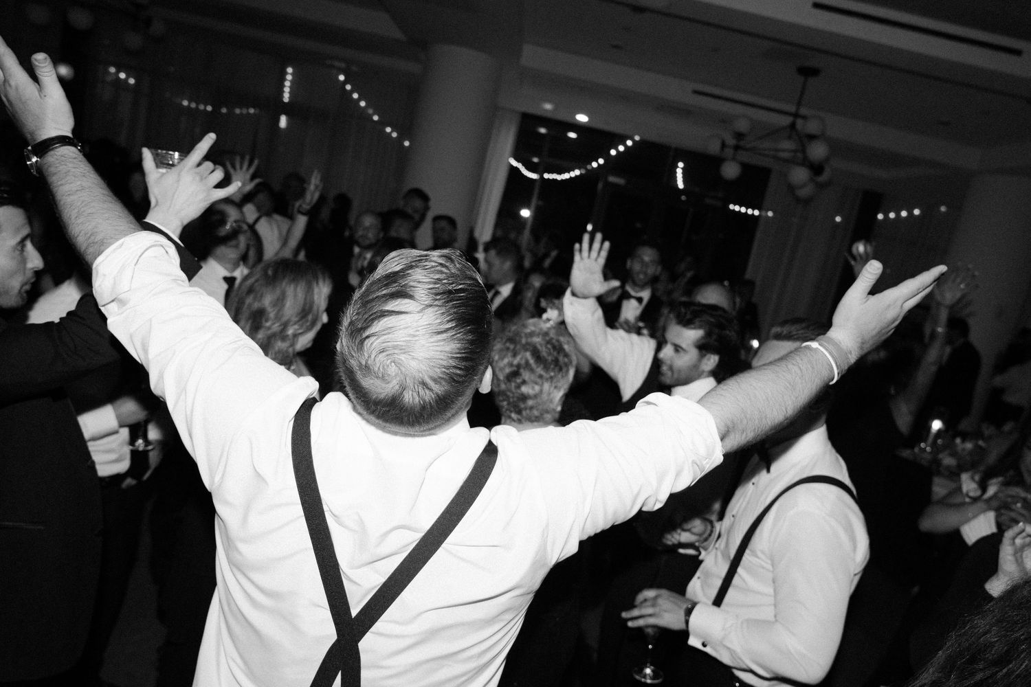 A guest with suspenders raises their arms in celebration while dancing at a wedding reception.