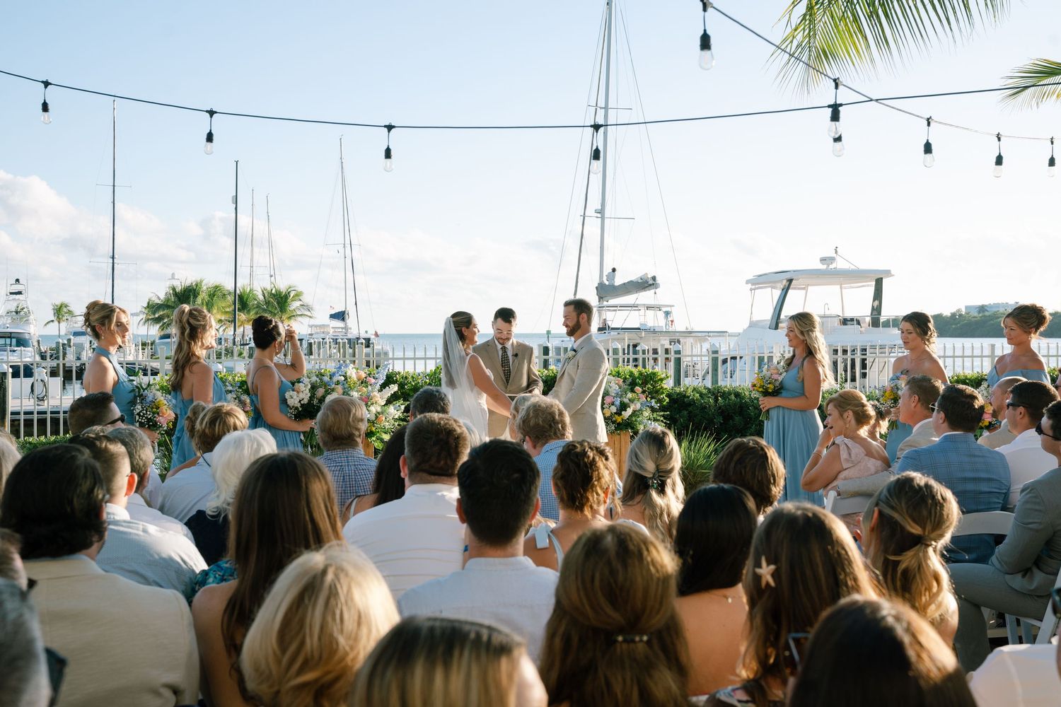 Wedding ceremony guests seated outdoors with string lights and palm trees overhead during sunset.