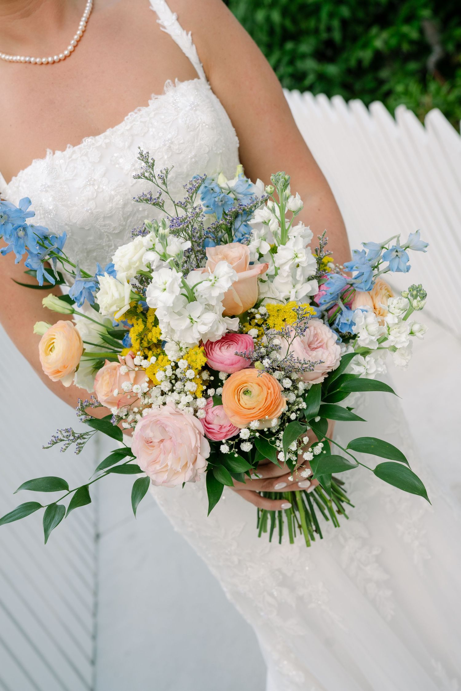 A beautiful bridal bouquet featuring peach roses, blue delphiniums, white baby's breath and greenery against a white dress.