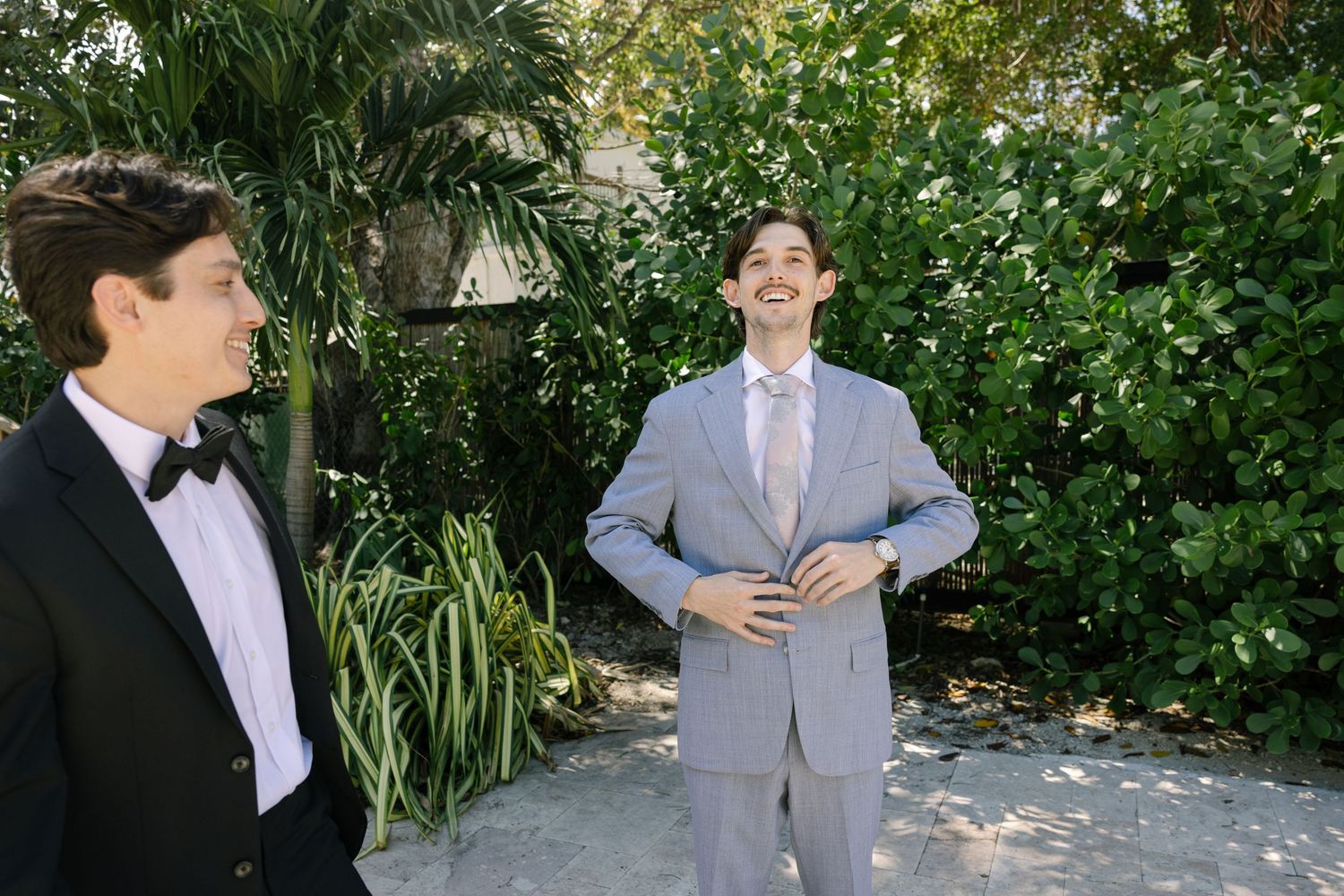 Two men in formal attire stand in a garden with lush greenery in the background.