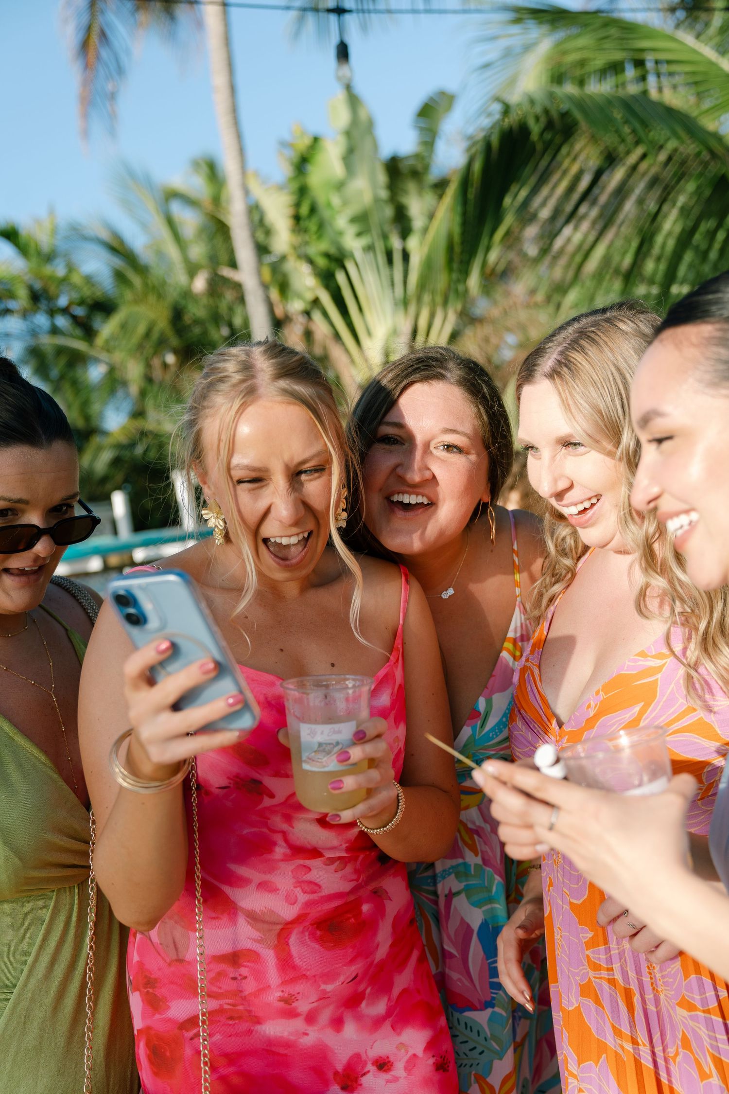 A group of women in colorful dresses and swimsuits taking a selfie together in a tropical setting.