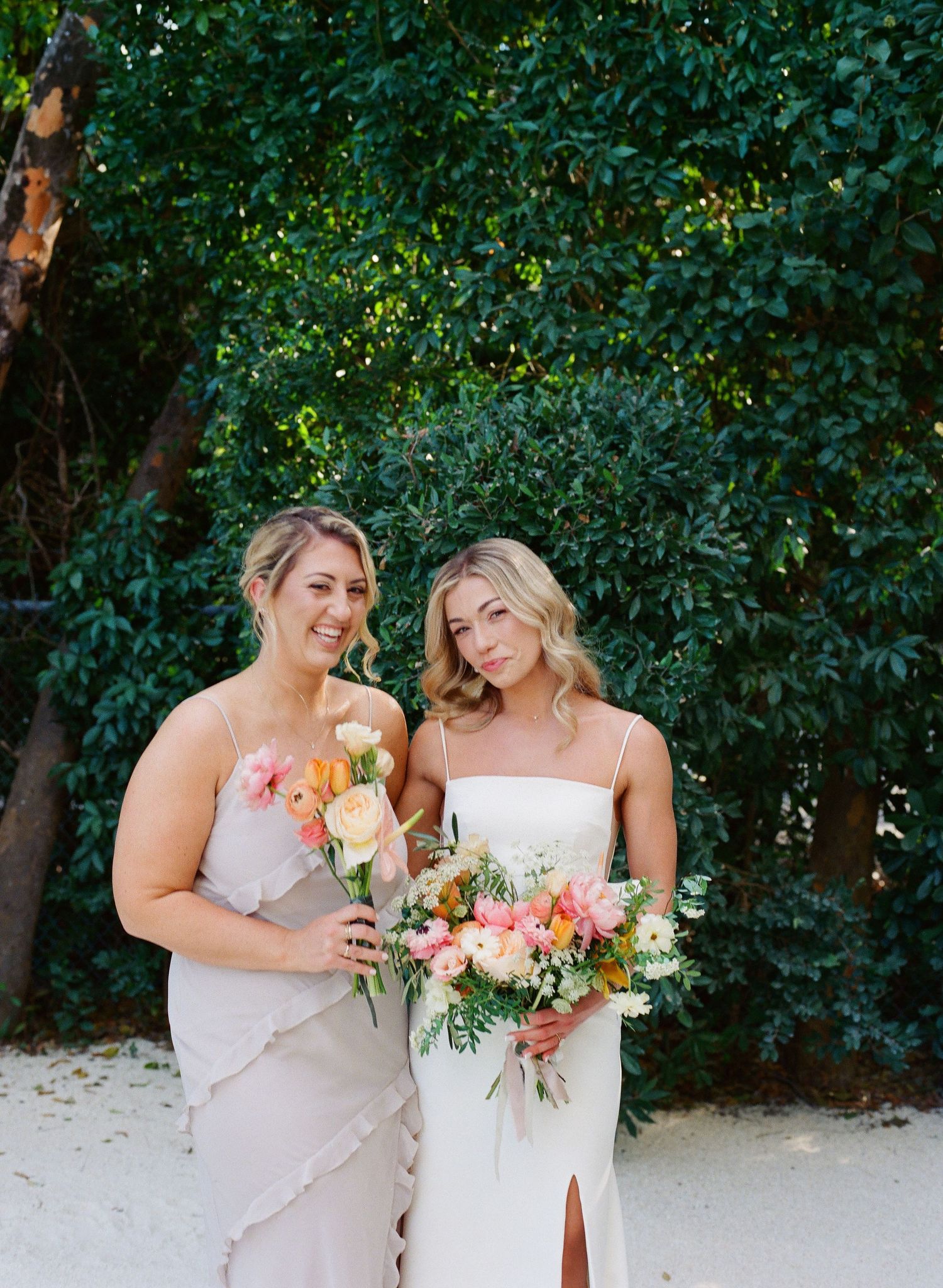 Bride and bridesmaid share joyful moments in a lush garden setting while holding coral and white floral bouquets.