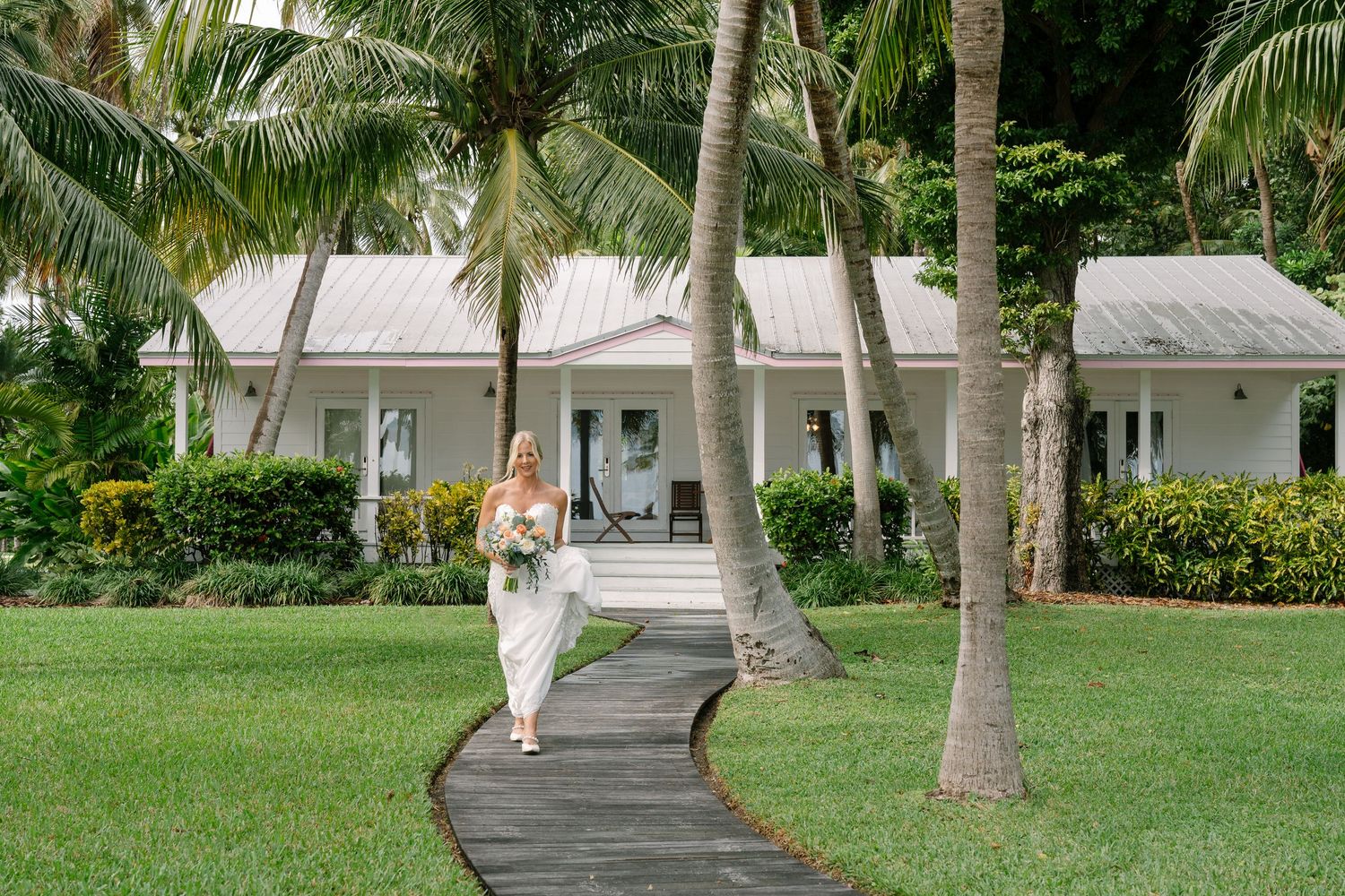 Winding pathway leads to white tropical bungalows surrounded by palm trees and lush gardens in a resort setting.