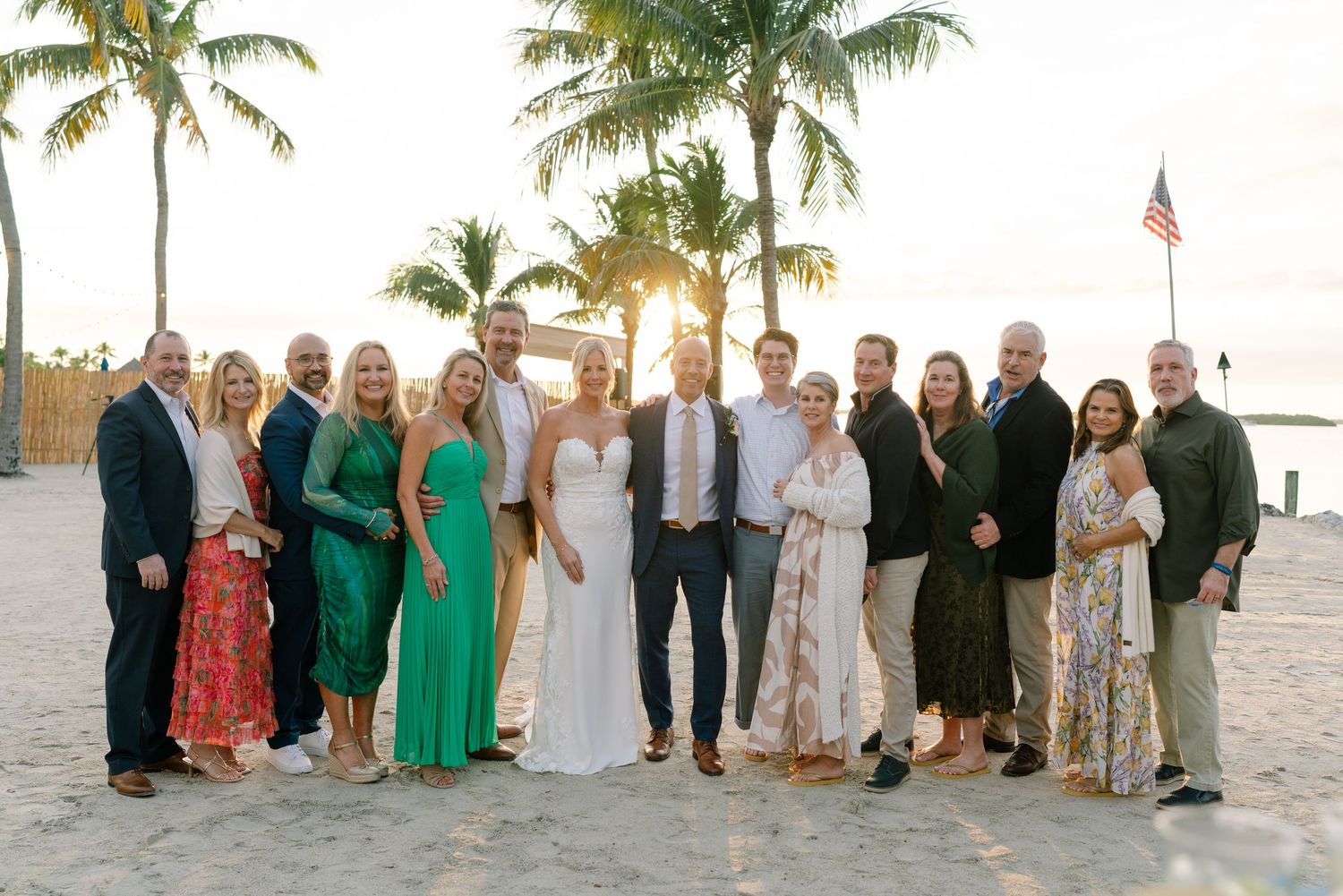 Large wedding party group photo on a beach at sunset with palm trees.