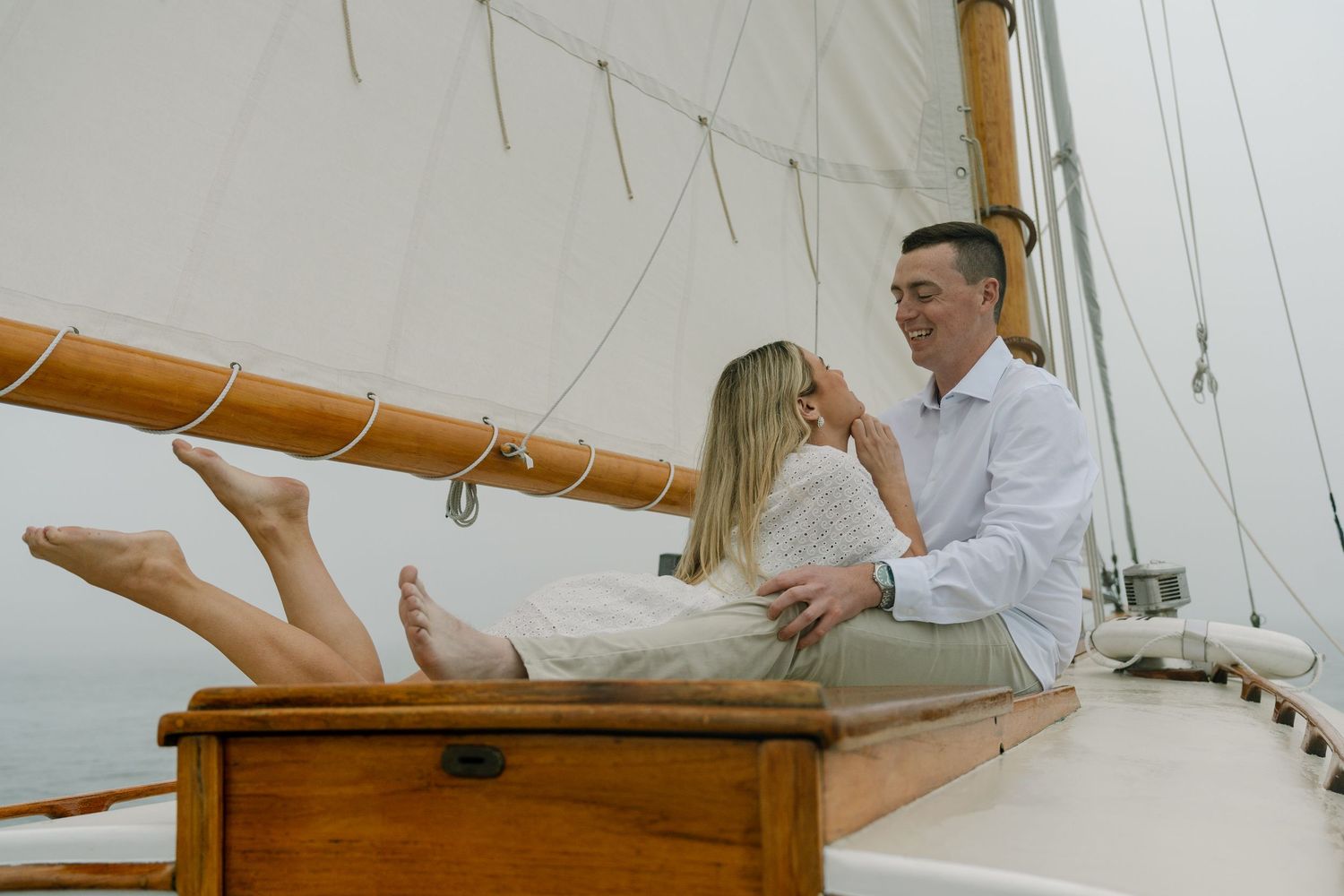 Couple relaxing on a classic wooden sailboat deck, sharing a romantic moment on a misty day at sea.