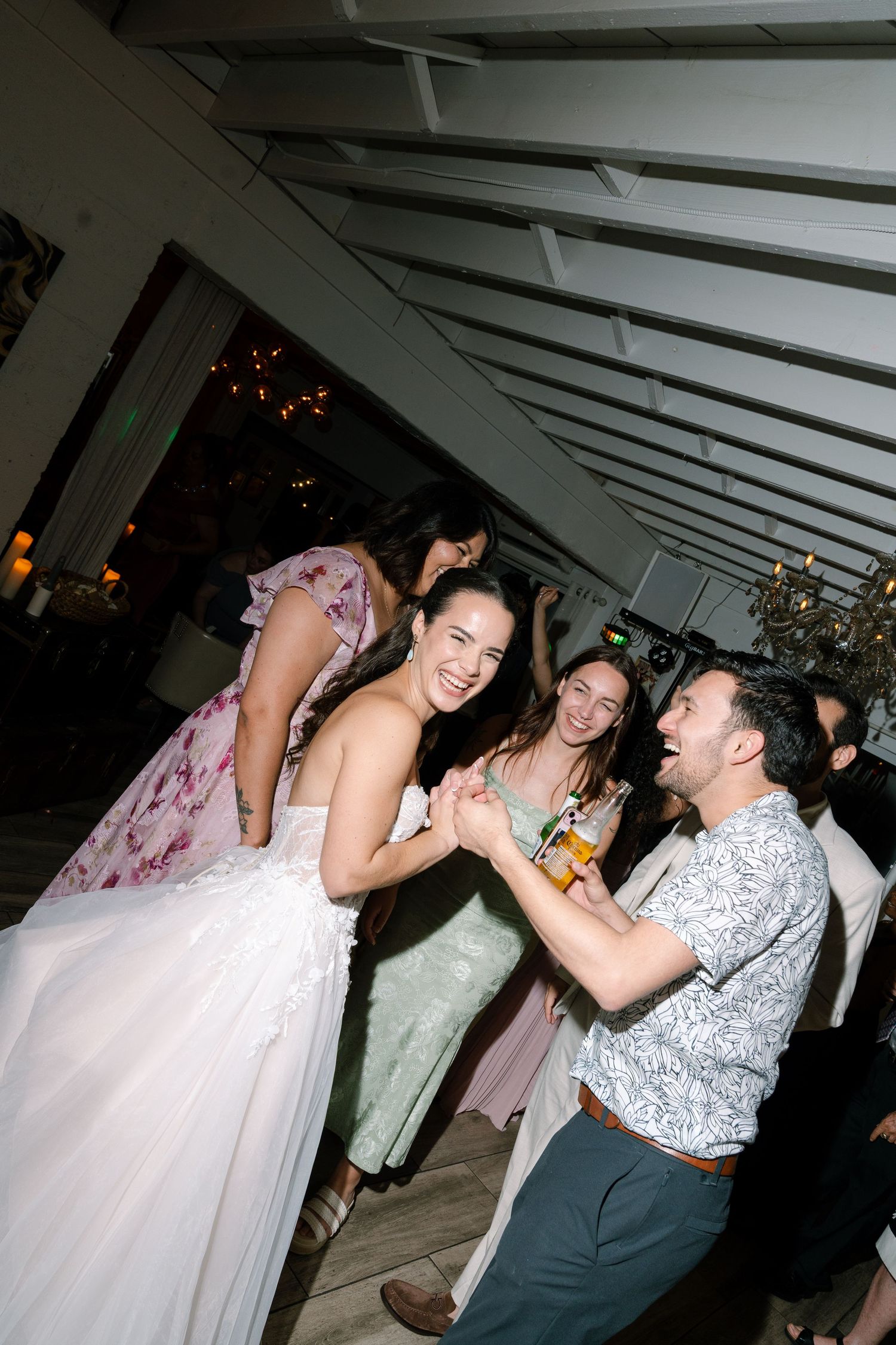 A bride in a white gown dances joyfully with a man kneeling at a wedding reception party.