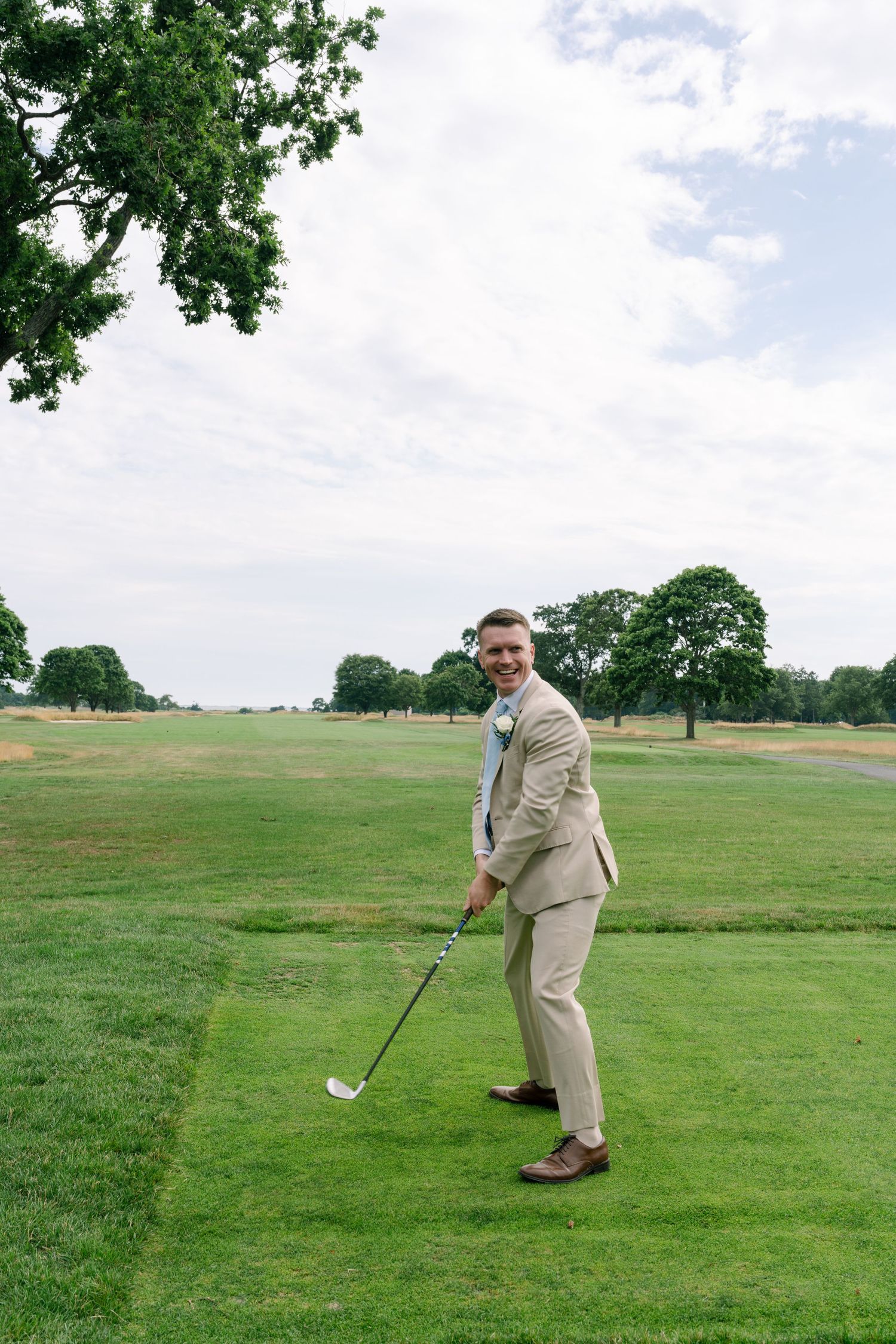 Smiling groom in beige suit holding golf club on lush green golf course on wedding day.