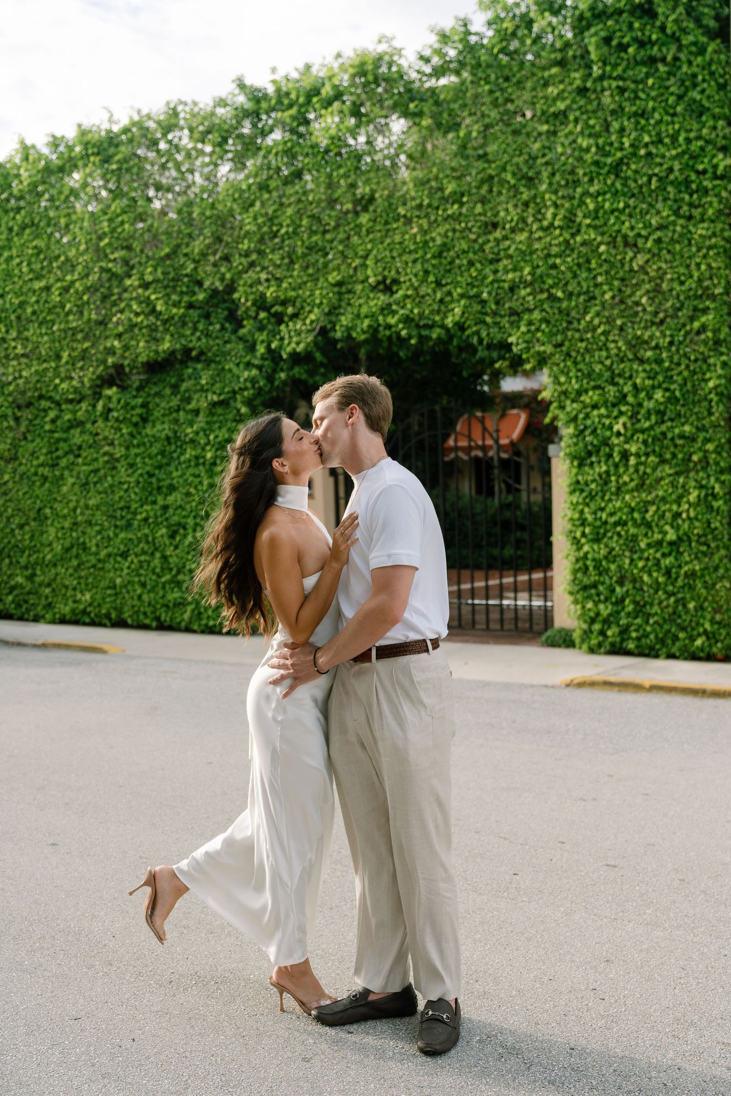 Couple sharing a romantic kiss outdoors, dressed in white and beige, in front of a lush ivy-covered wall.