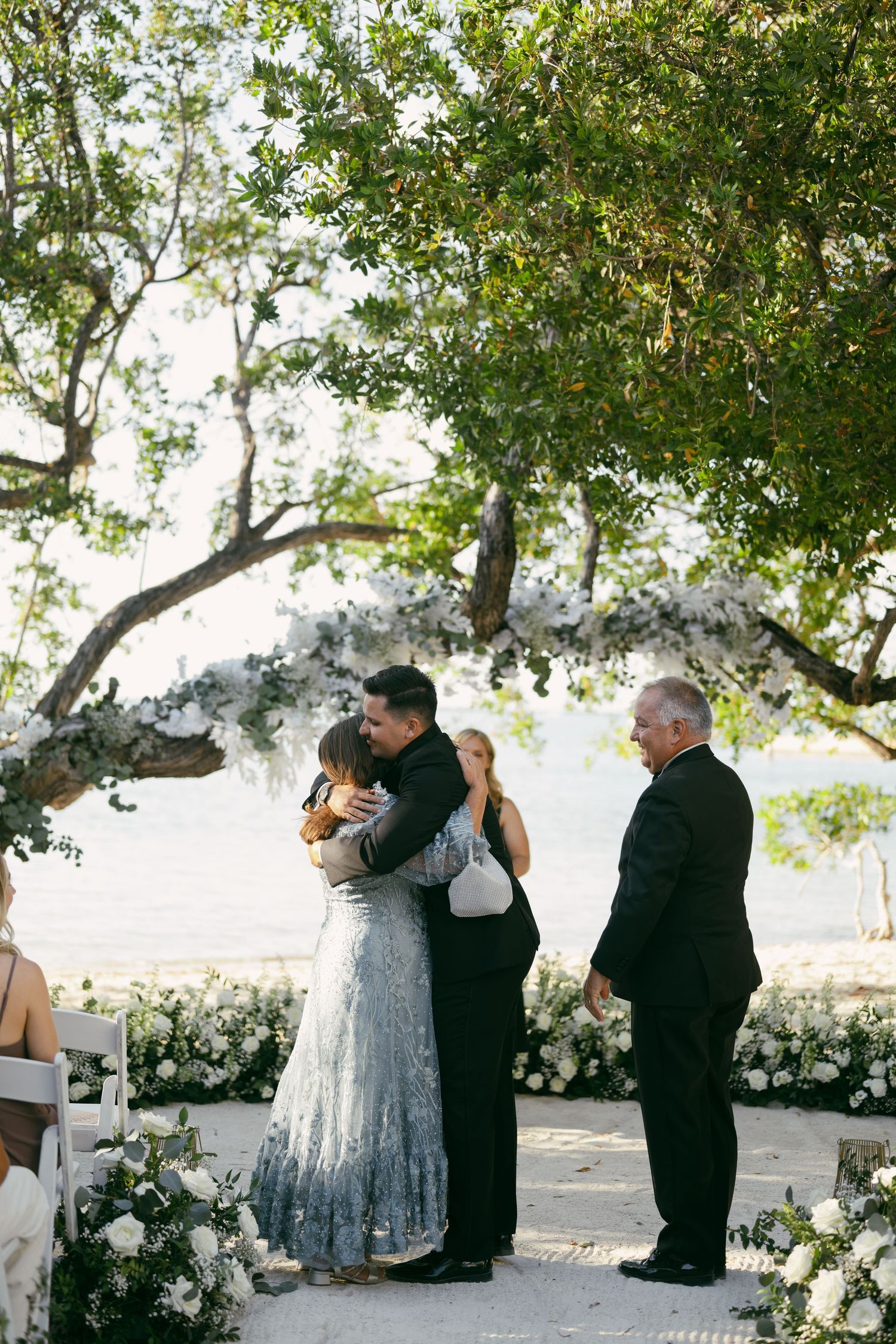 Three moments during an outdoor wedding ceremony under trees surrounded by white floral decorations and a small gathering.