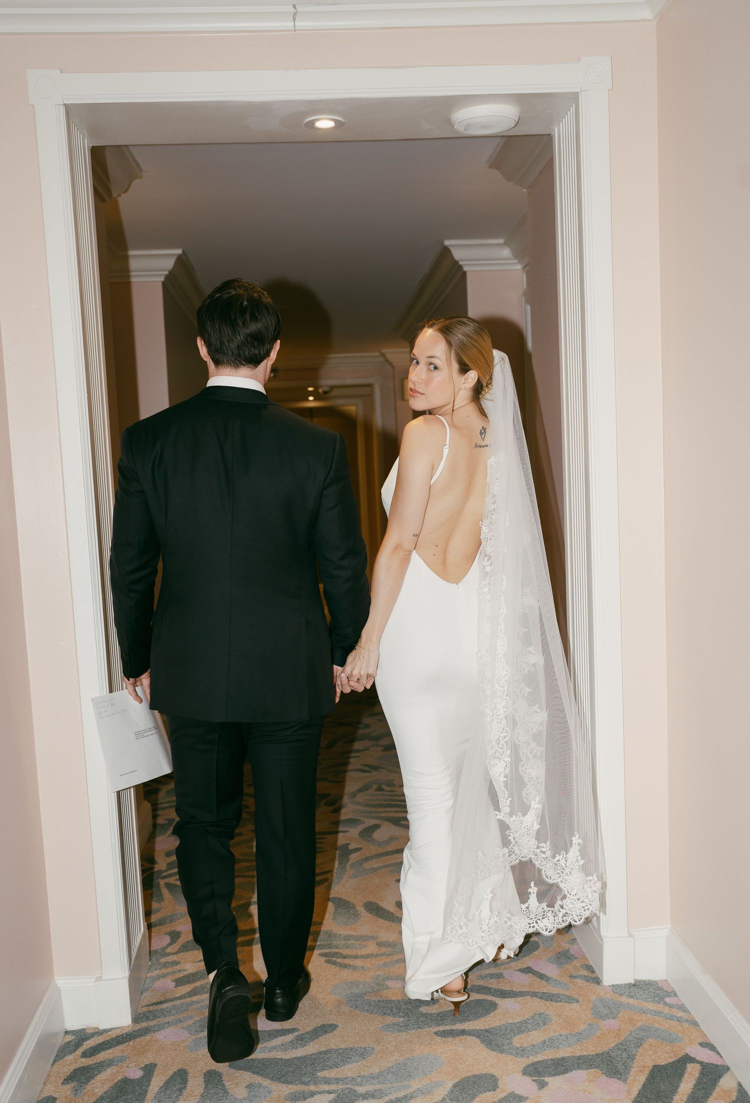A couple in wedding attire walking down a carpeted hallway, holding hands, with the bride looking over her shoulder.