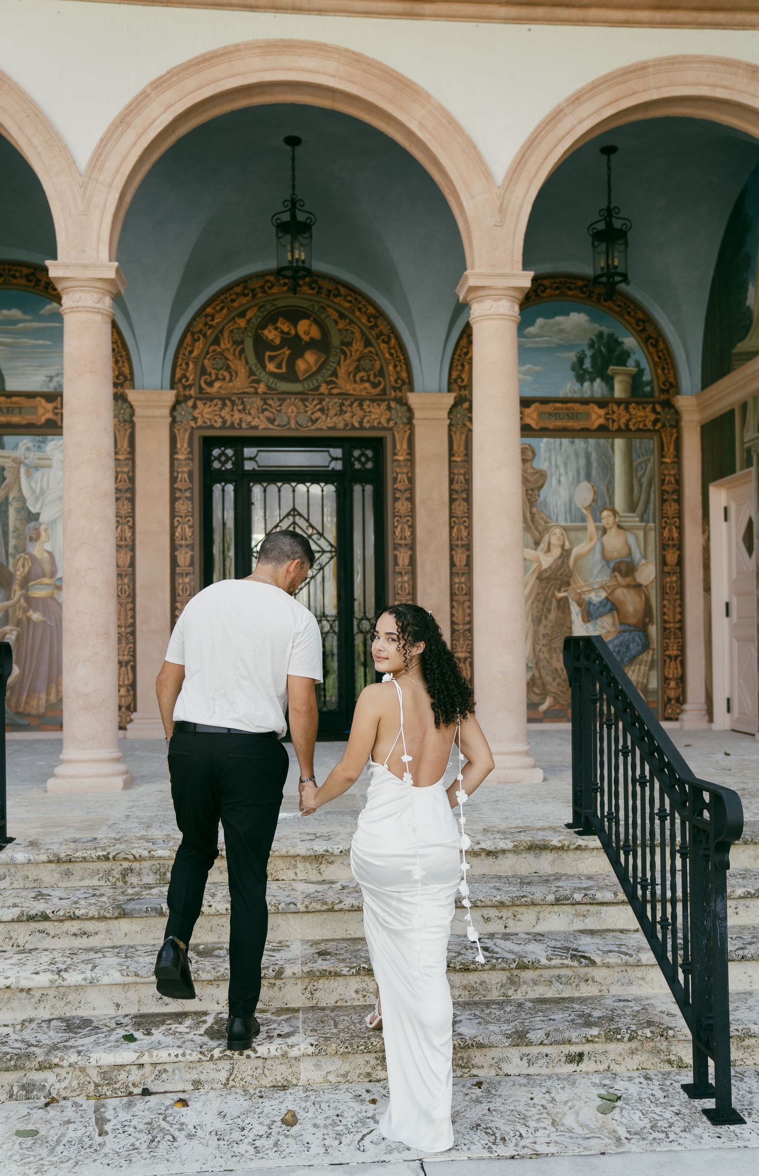 A couple ascends stone steps beneath ornate arched columns and painted murals at a historic Mediterranean-style venue.