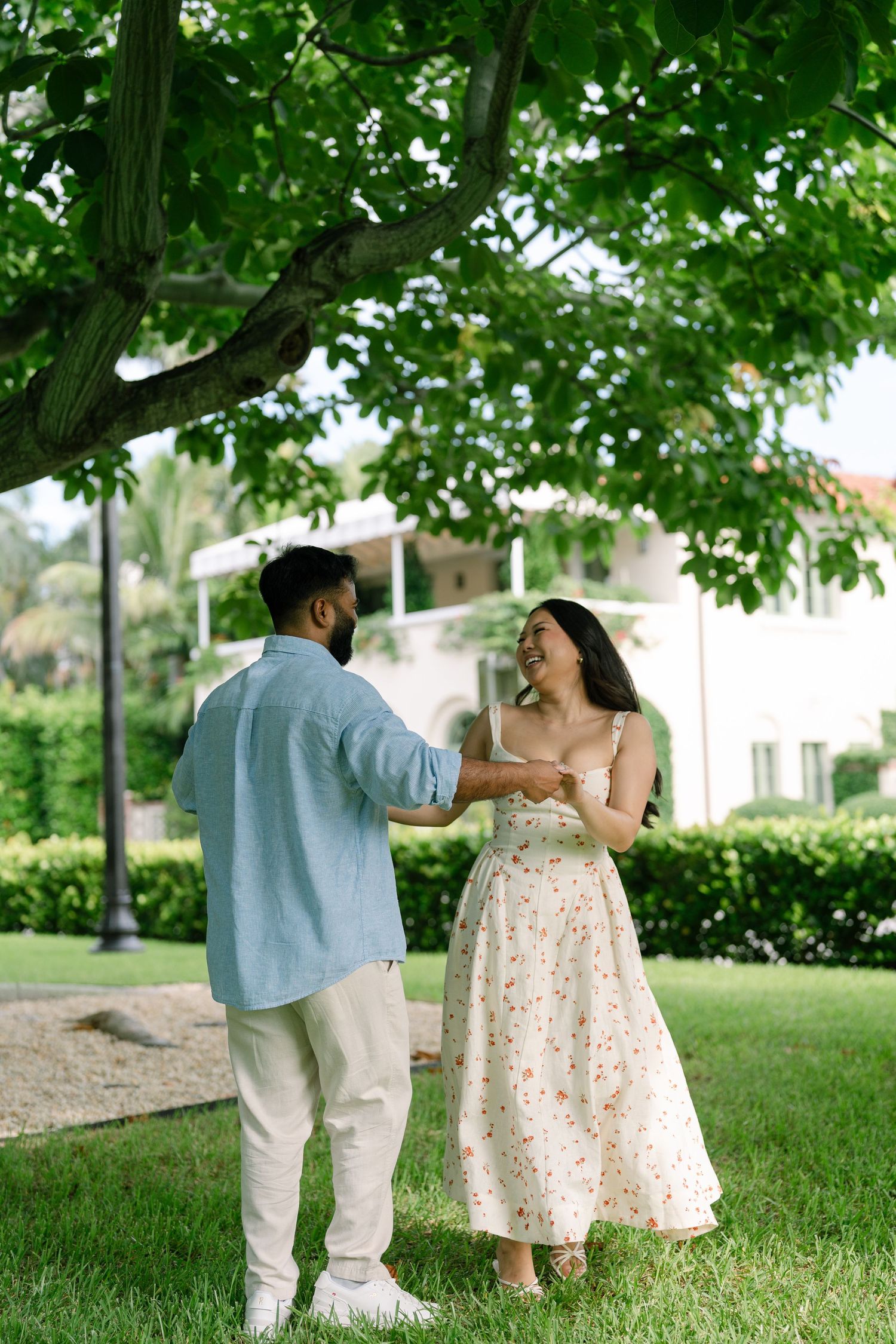 Couple holding hands and laughing together on green lawn under leafy tree near white building on sunny day.