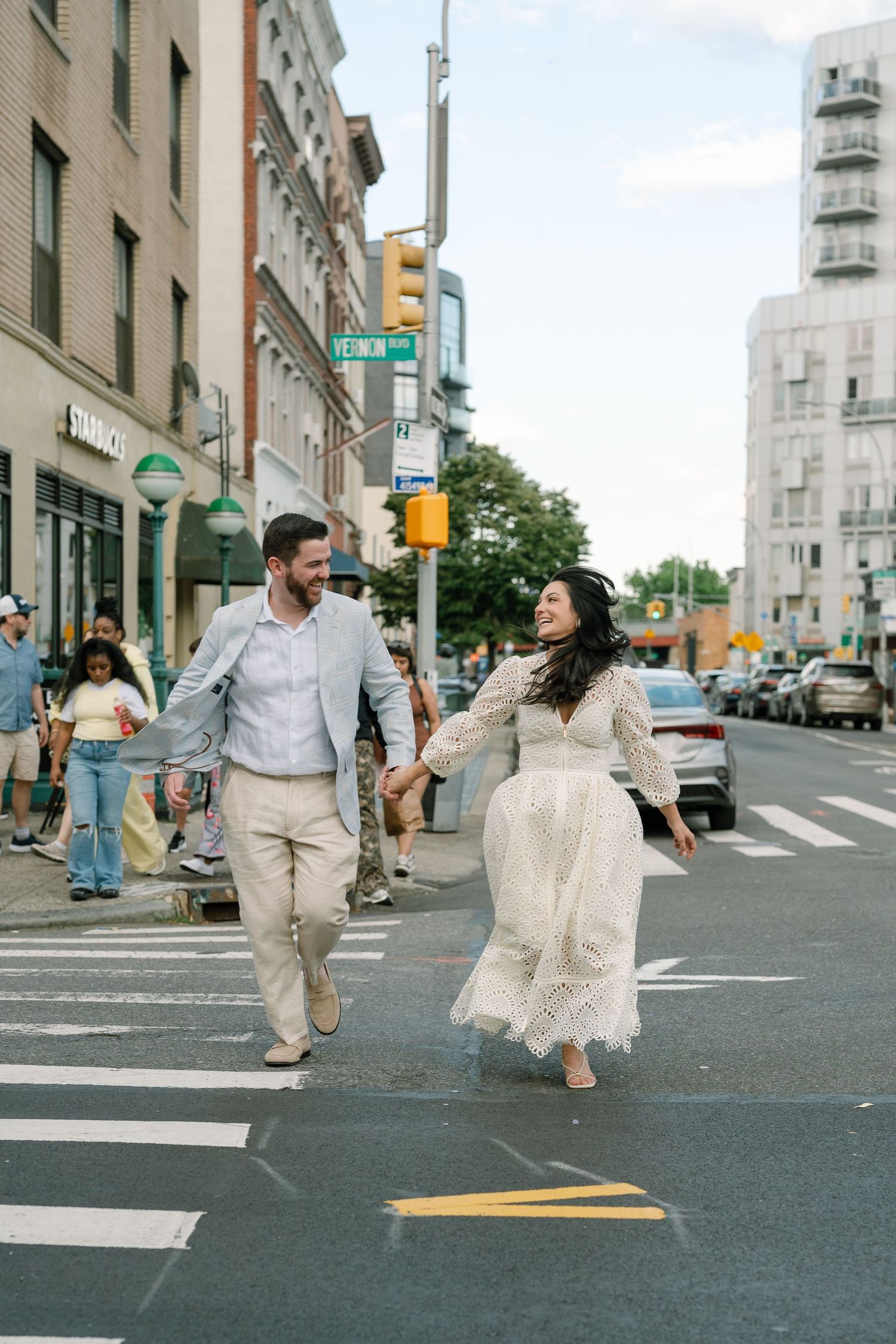A couple in wedding attire holds hands while crossing a busy urban street intersection, smiling joyfully.