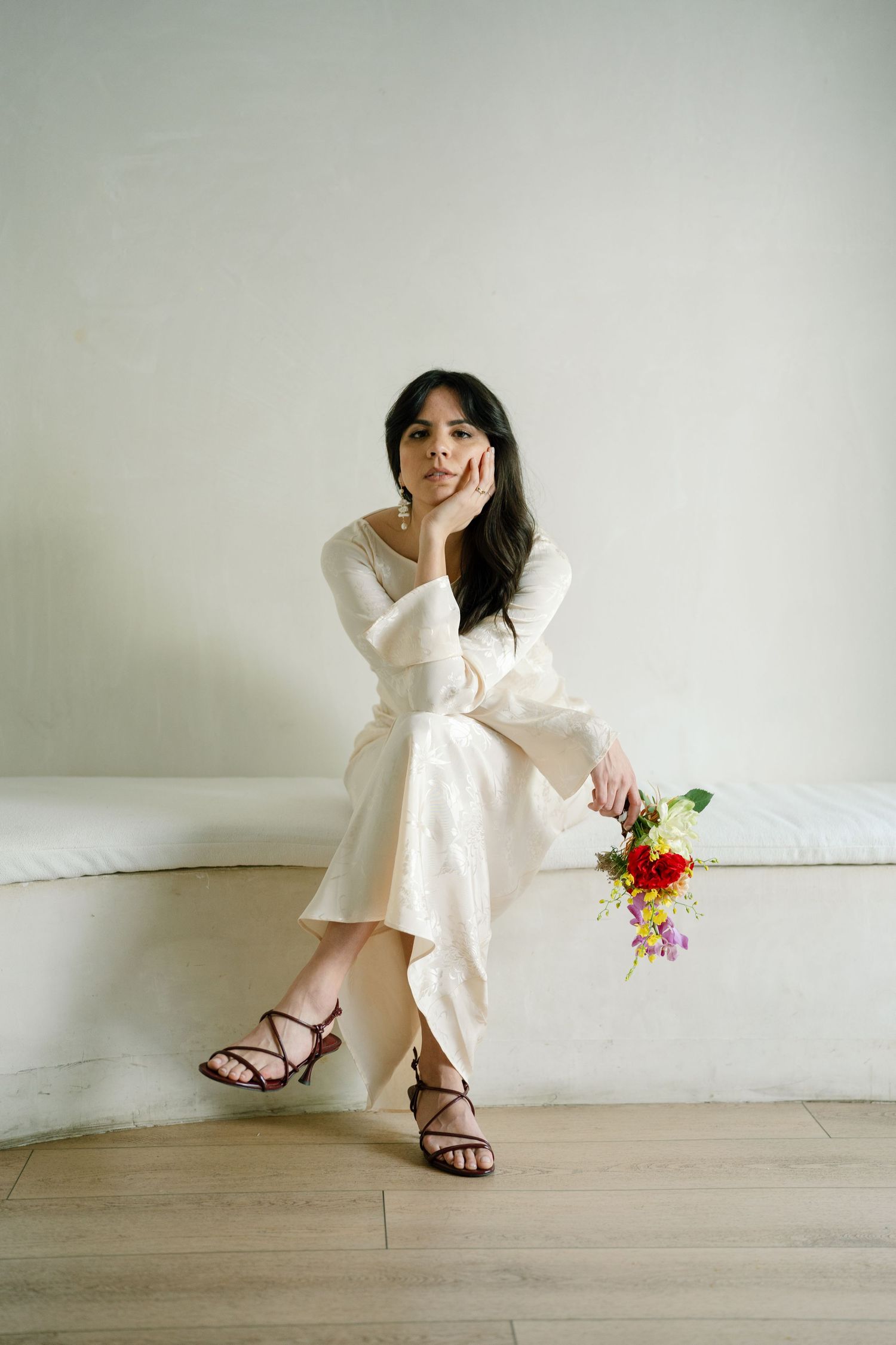 A person in a white dress sits on a bench holding colorful flowers and smiling.