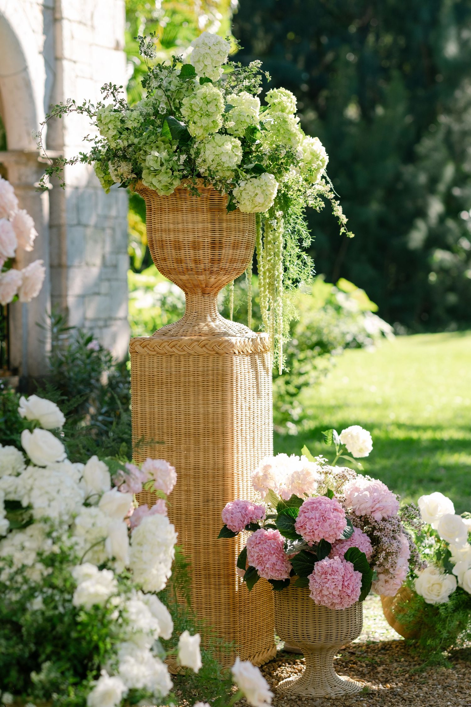 Tall wicker urns filled with white hydrangeas and pink blooms decorate an outdoor garden wedding ceremony.