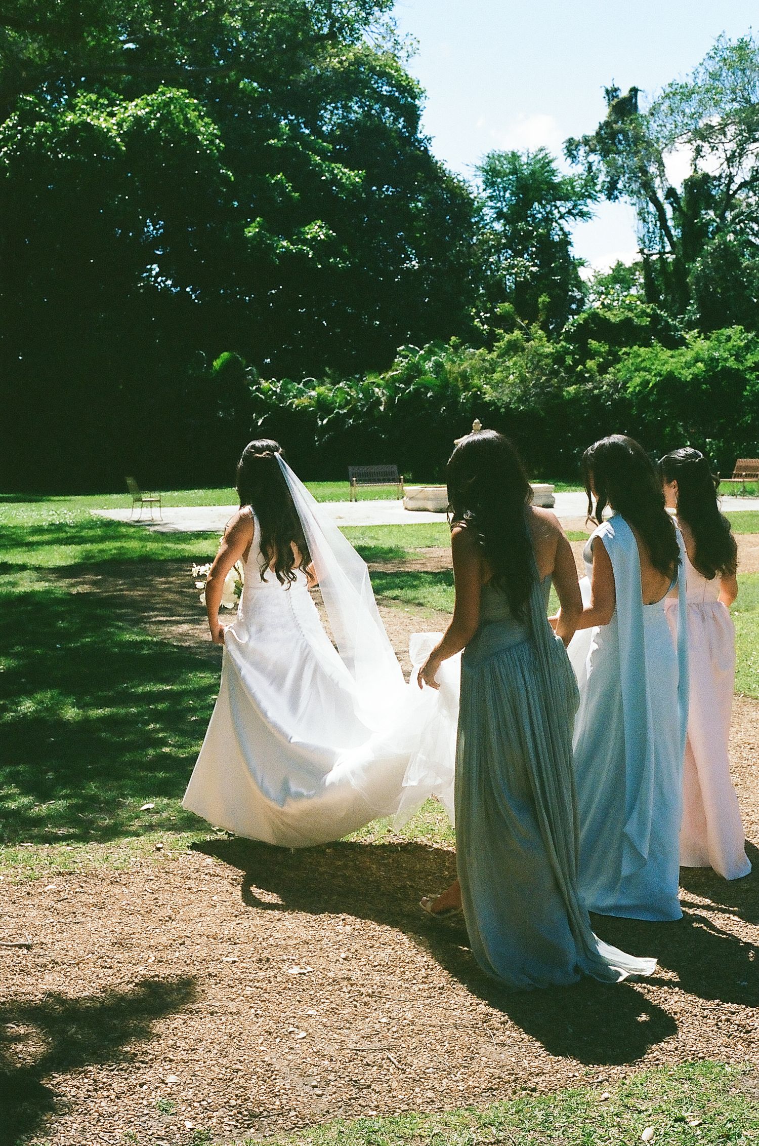 Bride in white gown and veil walks with bridesmaids in pastel dresses through a sunny park setting.