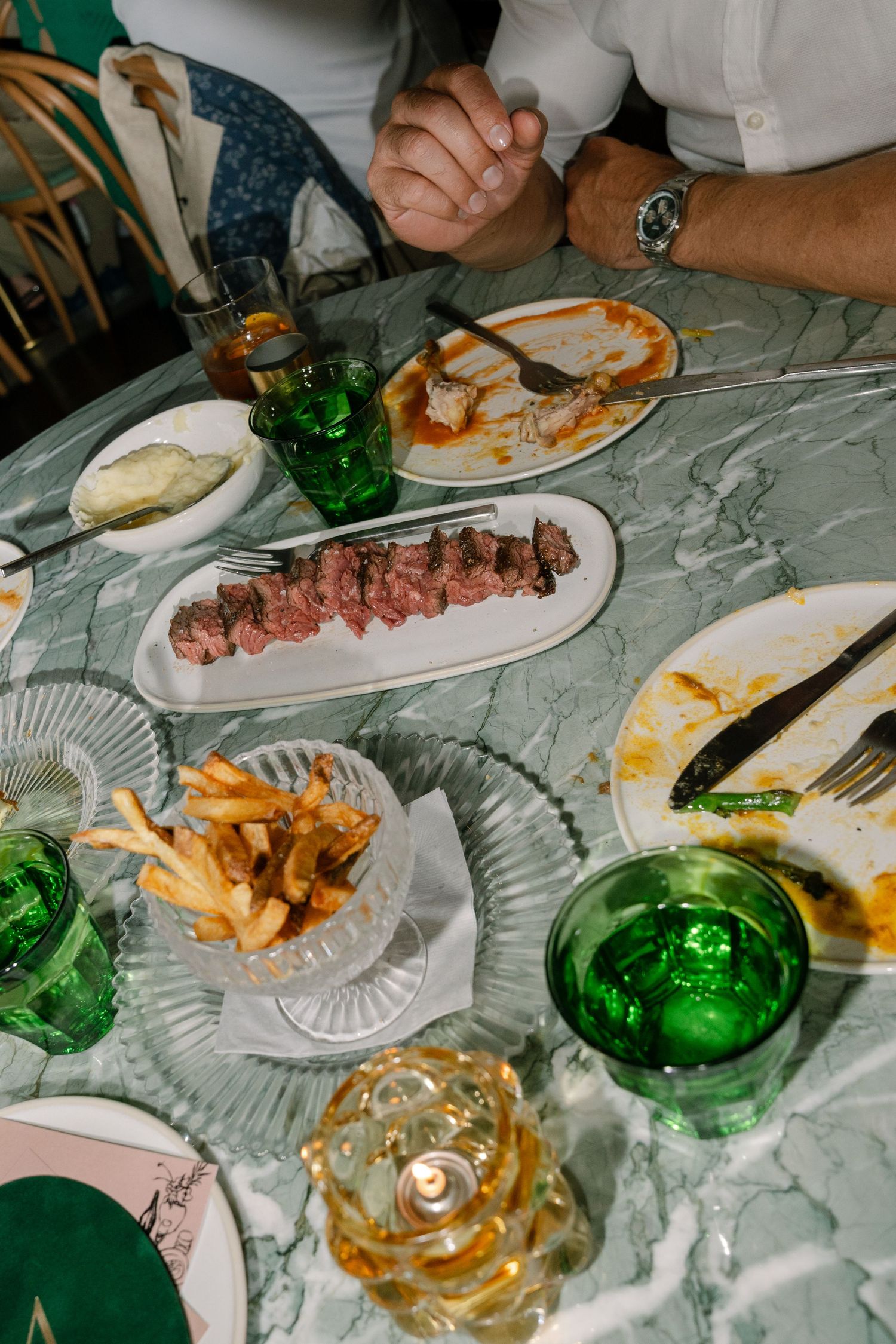 Dinner table spread with plates of grilled meats, sides, and green glass bottles on a white tablecloth.
