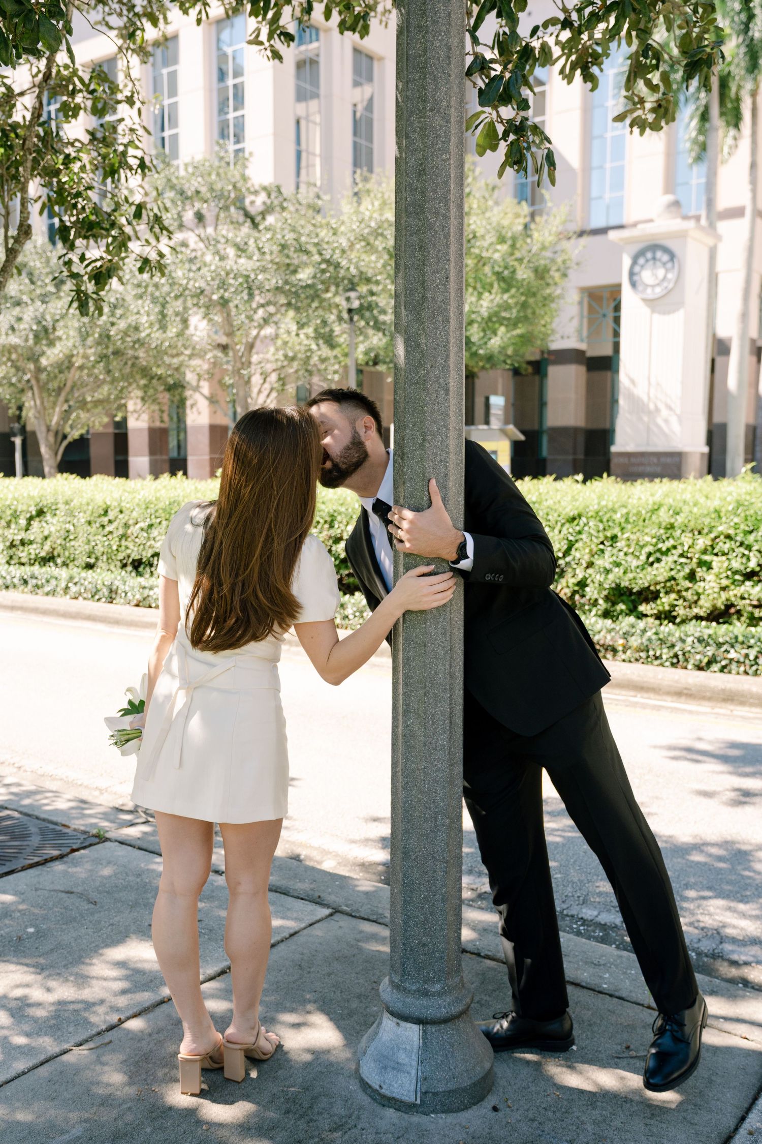 Playful couple poses around a concrete pillar in an outdoor setting with greenery.