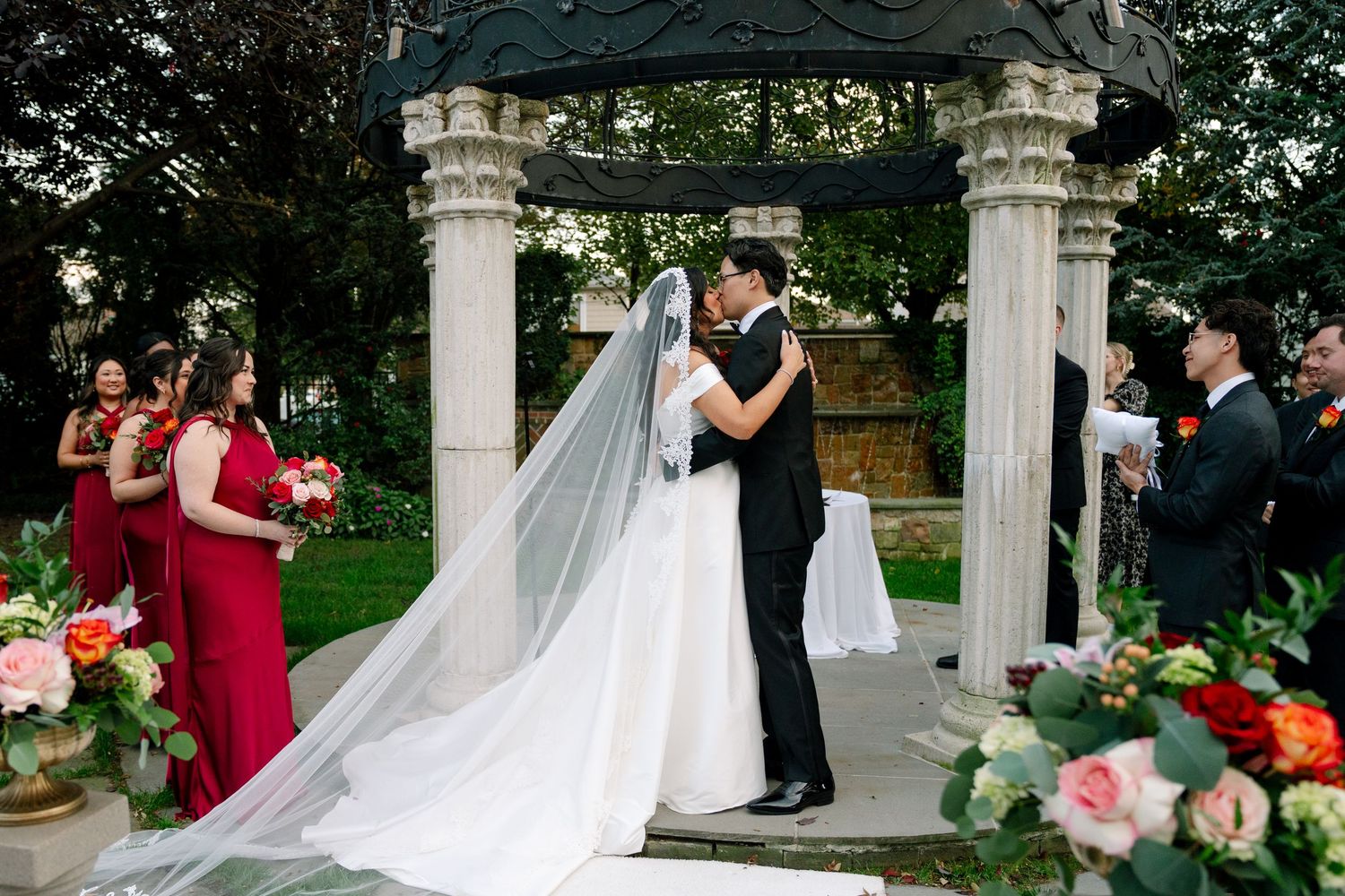 Outdoor wedding ceremony under white pillared gazebo with pink floral decorations.