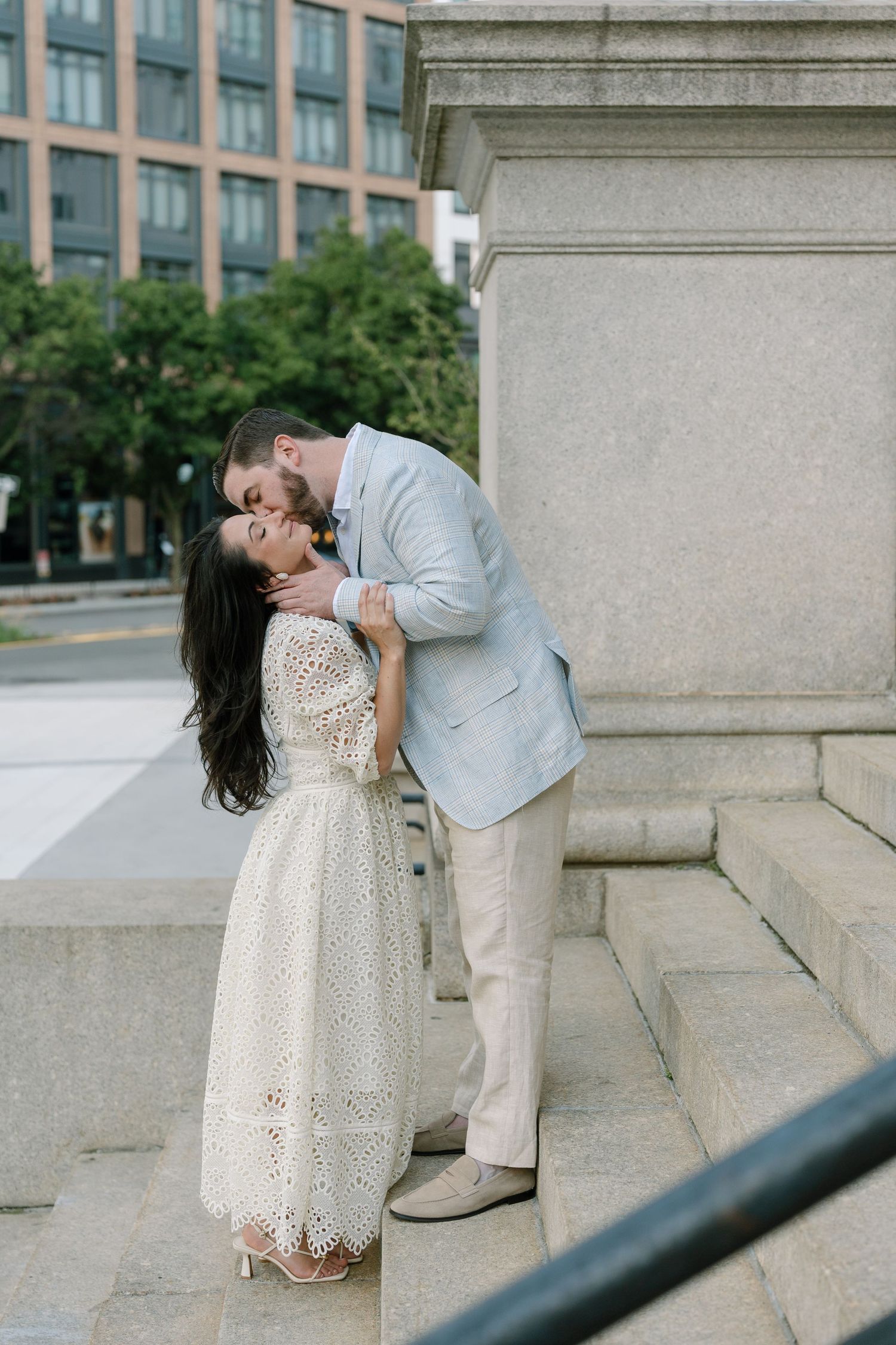 Couple sharing a romantic kiss on stone steps near a city monument, woman in white lace dress, man in light blue suit.