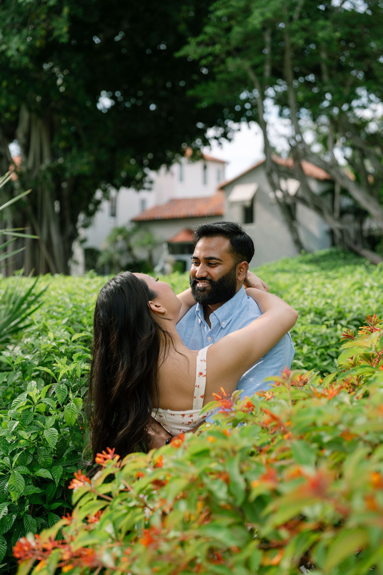 A smiling couple embraces romantically among lush green plants and orange flowers in a tropical garden setting.