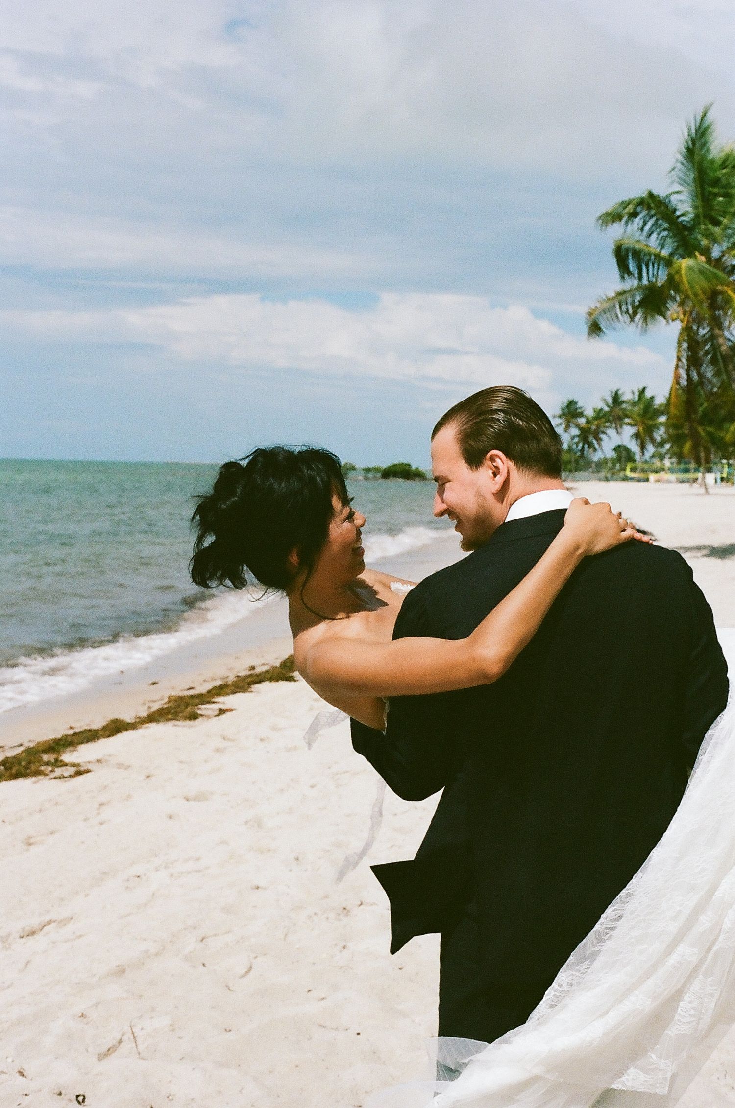 Bride and groom share a romantic embrace on a tropical beach with palm trees and ocean in the background.