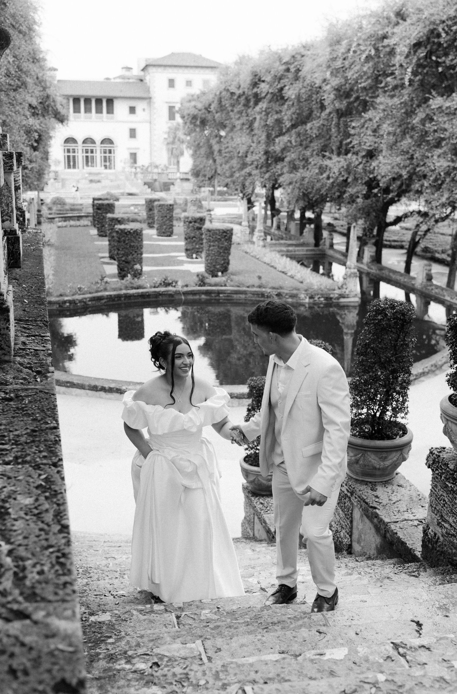 A black and white wedding photo showing a couple ascending stone steps alongside a reflecting pool at an ornate villa.