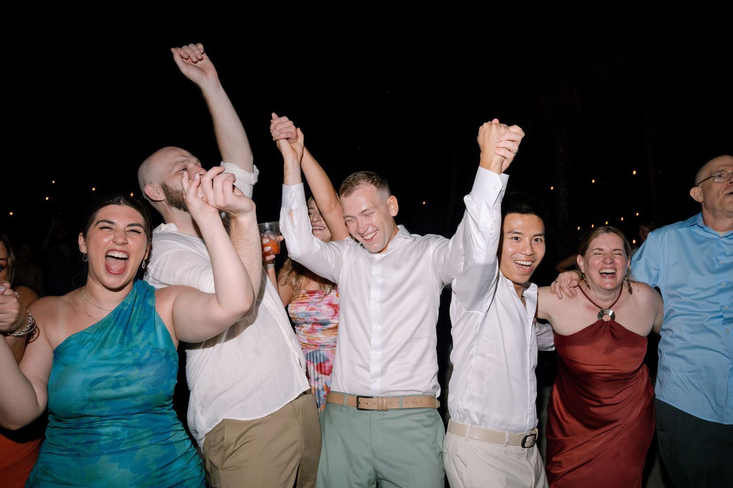 People dancing and celebrating with raised arms on a dark dance floor at a party or reception.