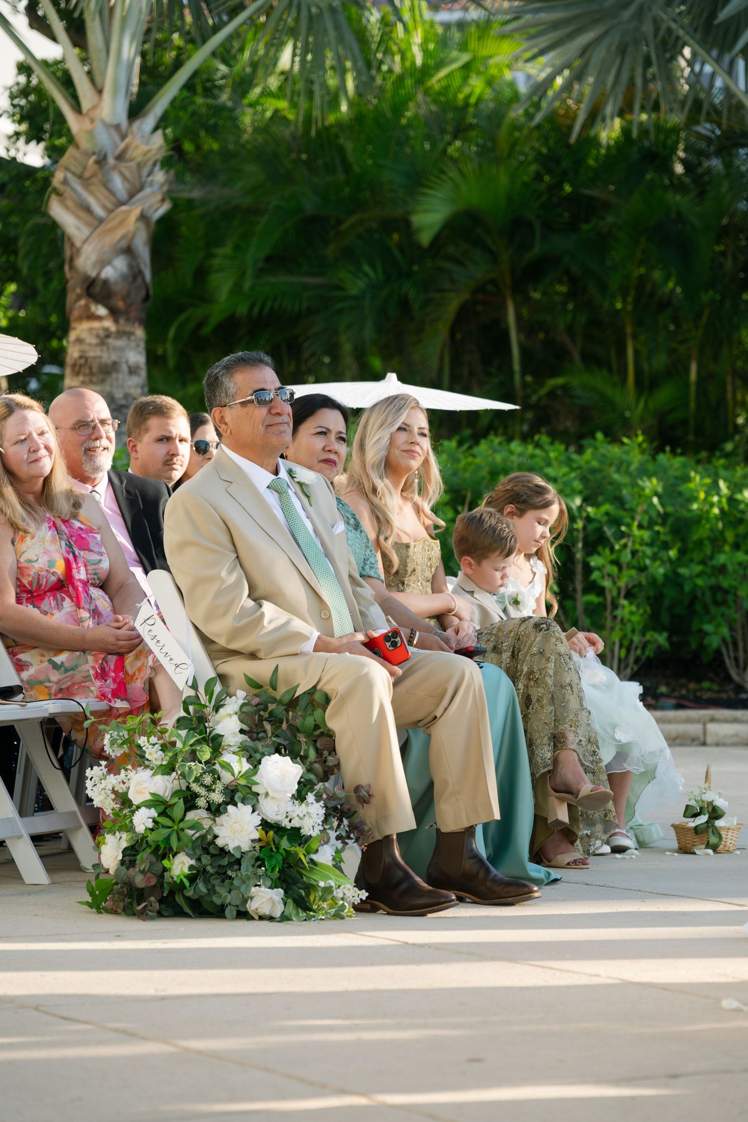 Wedding guests seated outdoors in formal attire with tropical palm trees in the background.