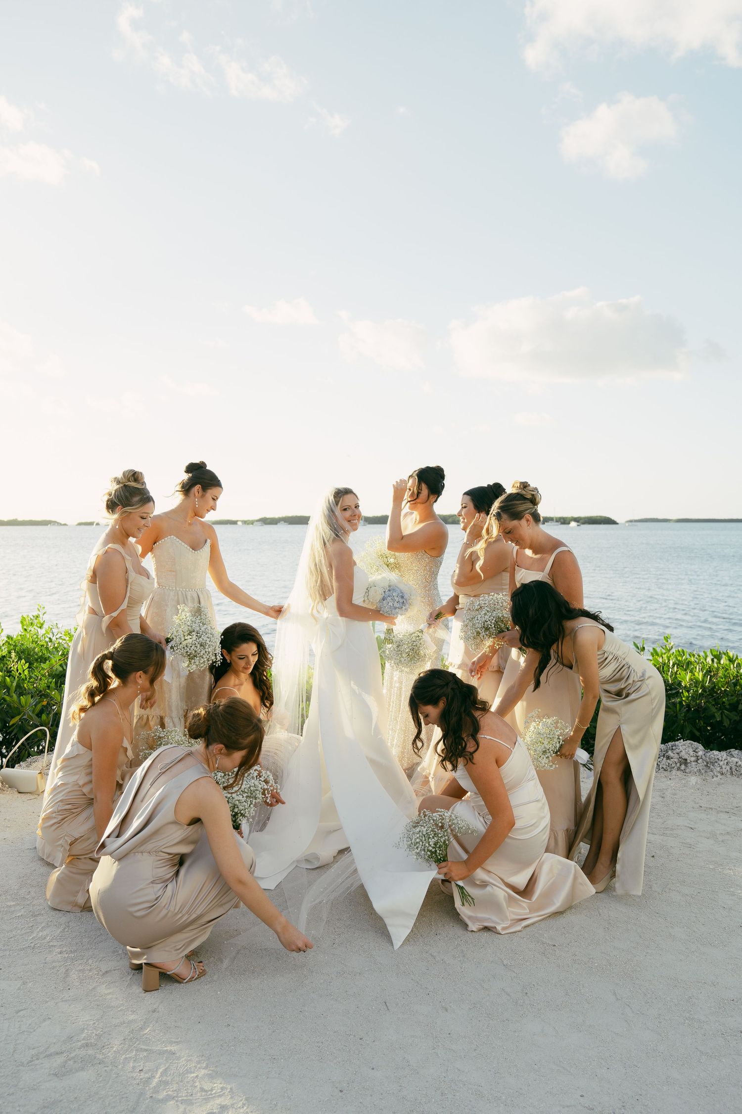 Bridesmaids in blush dresses gather around bride in white gown on a scenic beach with blue water and sky in background.