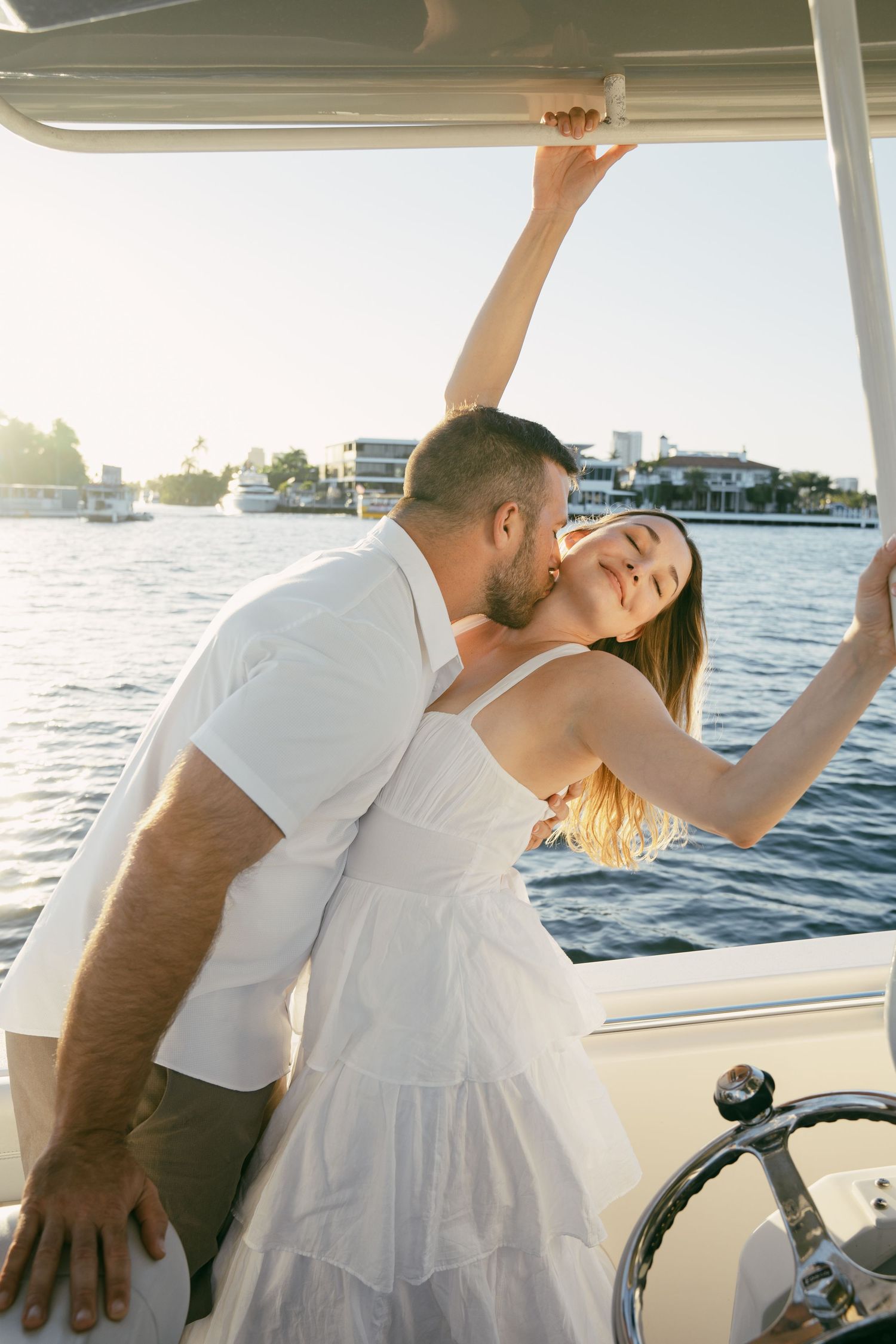 Couple in white outfits sharing a romantic moment on a boat during sunset with water and boats in background.