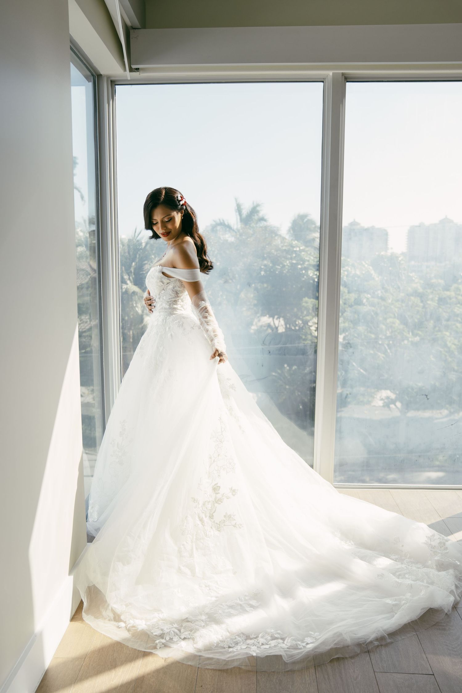 A flowing white wedding gown with a long train is backlit by sunlight streaming through large windows in an elegant room.