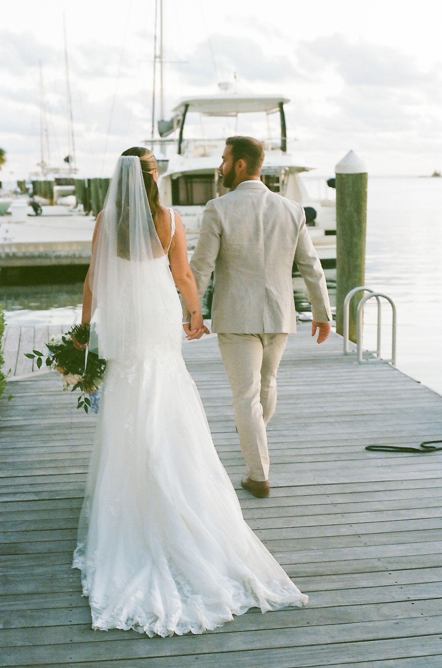 Wedding couple walking along wooden dock by waterfront in white dress and light colored suit.