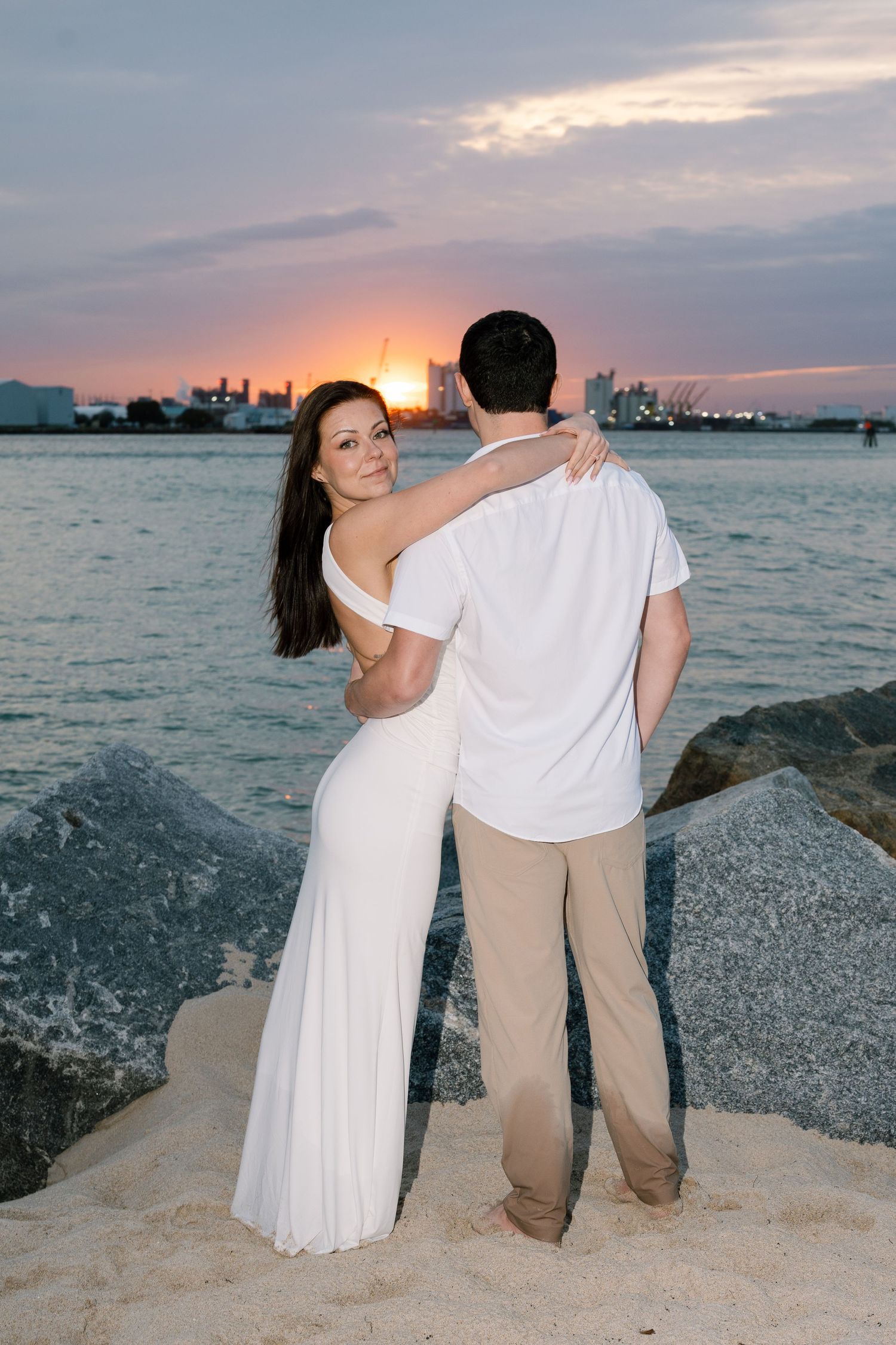 A couple sits together on rocky shoreline during sunset overlooking the ocean and city skyline.