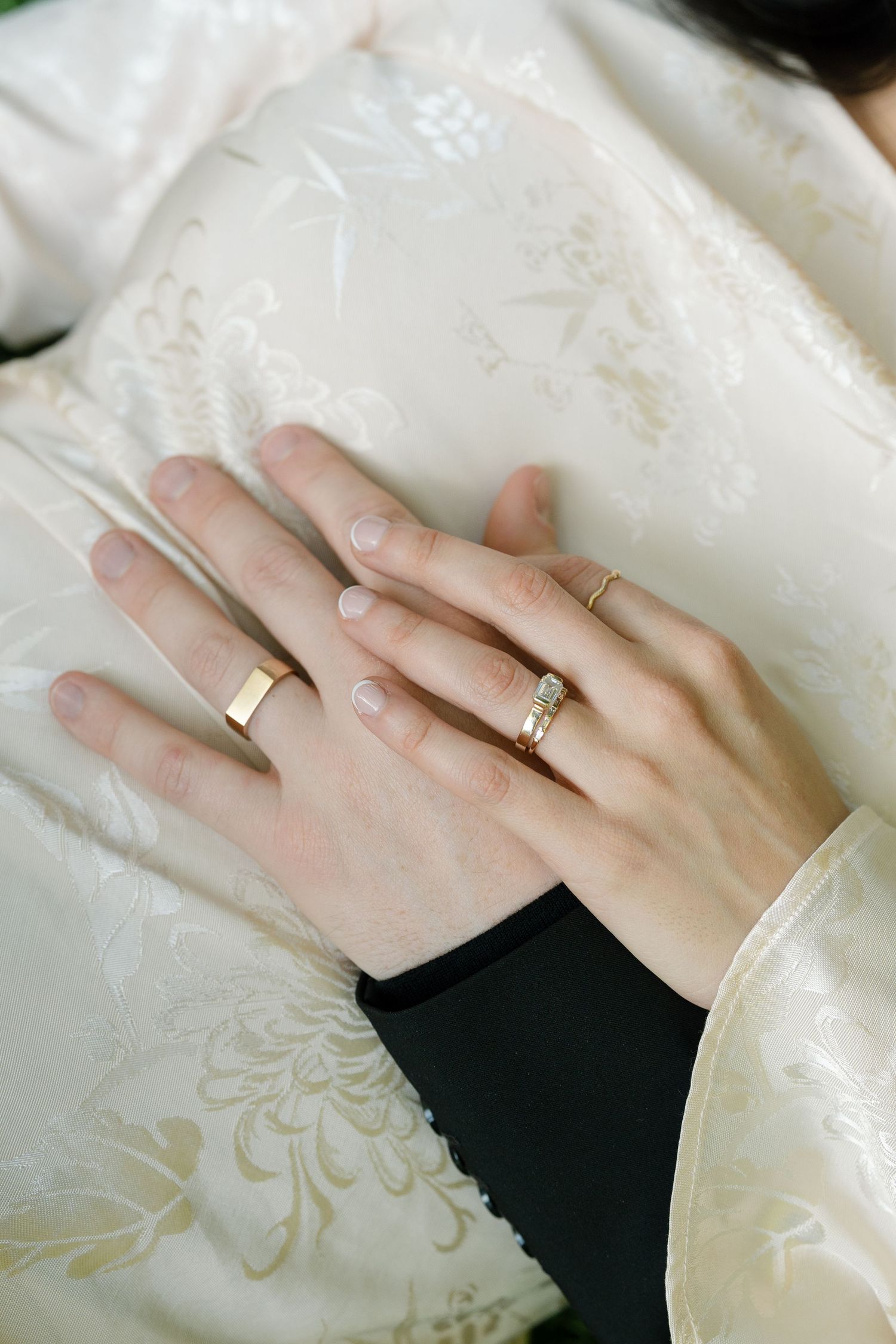 A hand wearing a gold wedding band rests against a white fabric with delicate floral patterns.