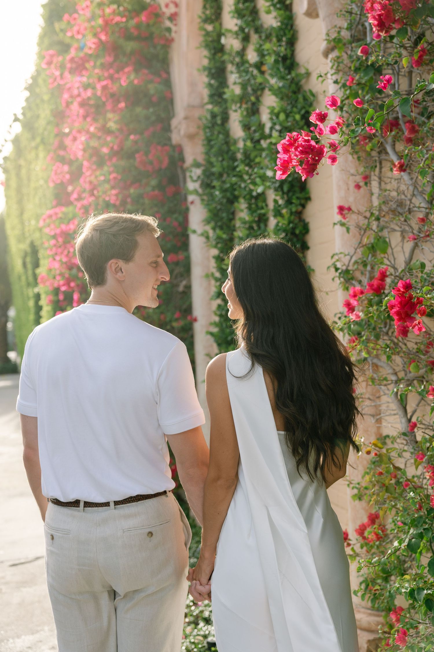 Couple holding hands walking by a flower-covered wall with pink bougainvillea in warm sunlight.