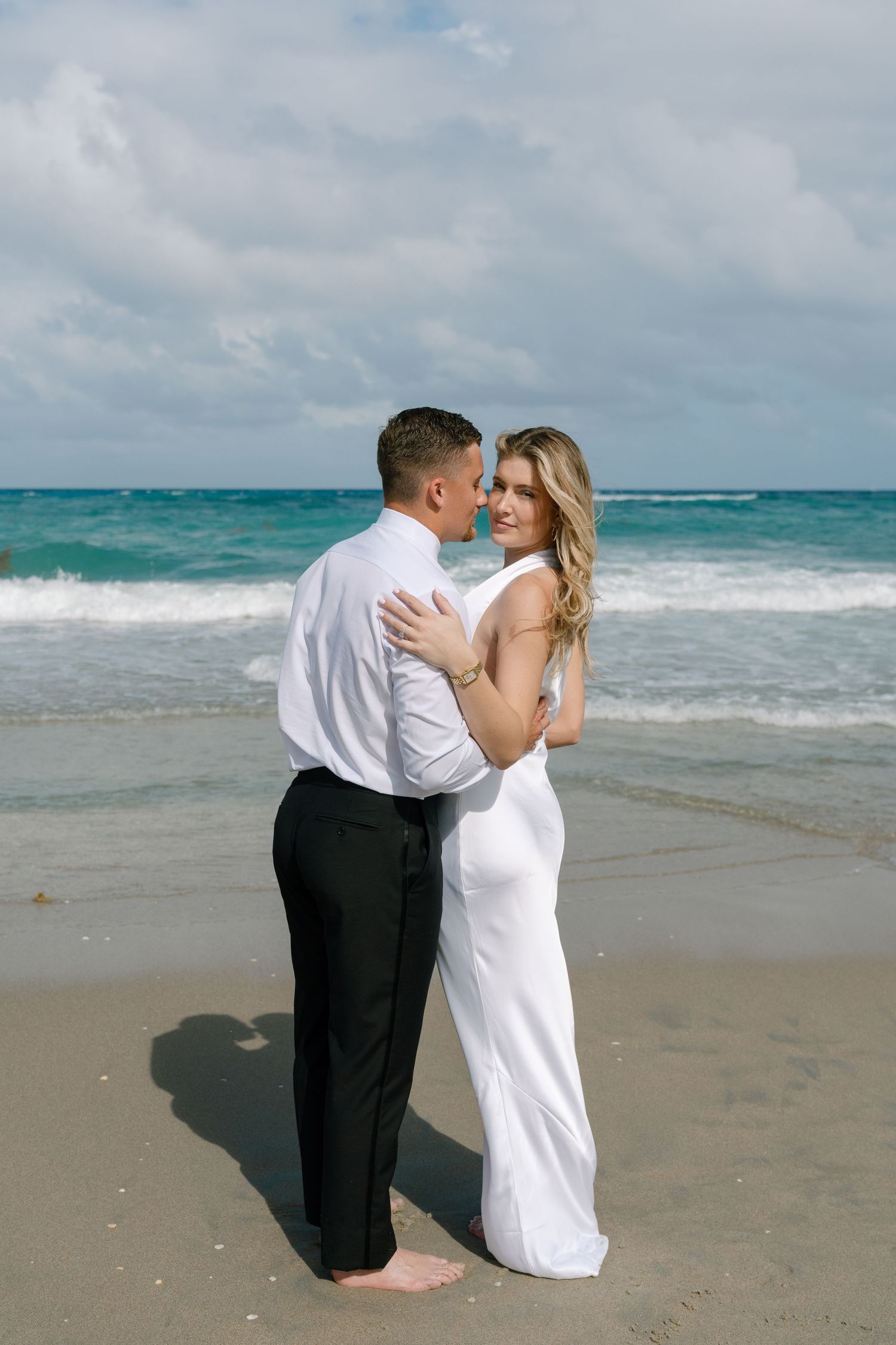 Bride in white gown and groom embracing romantically on sandy beach with turquoise ocean waves behind them.